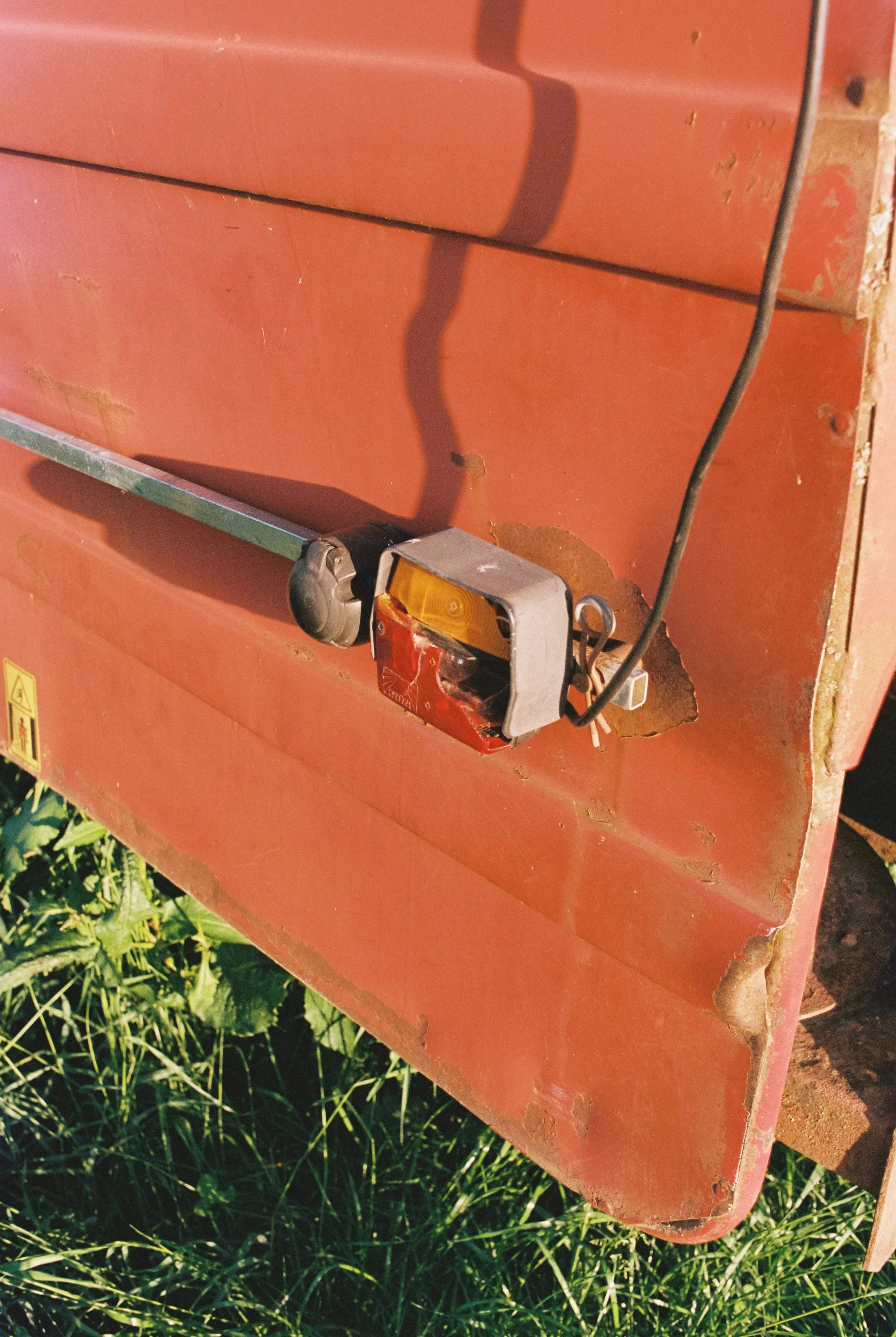 Ein älteres rotes landwirtschaftliches Fahrzeug mit einem defekten licht und angeschlossene Kabel, das auf grünem Gras steht. Geschossen auf Portra 400.