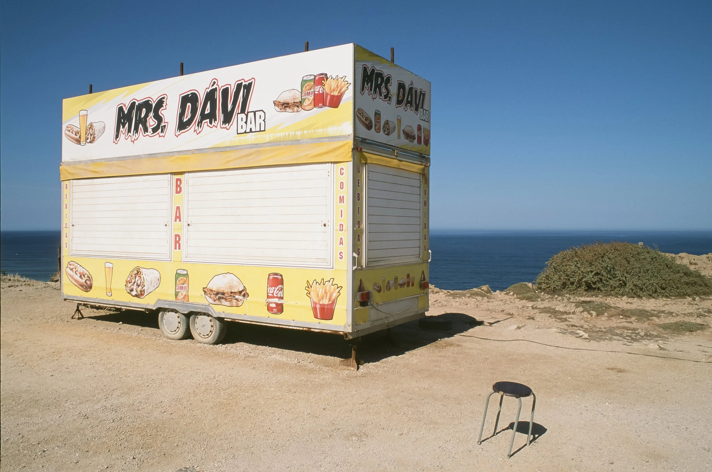 Gelber Imbisswagen mit dem Schriftzug 'Mrs. Dáv Bar' an einer Küste, mit Bildern von Fast Food und Getränken, Blick auf den Ozean. Portugal auf Fuji Provia 100f.
