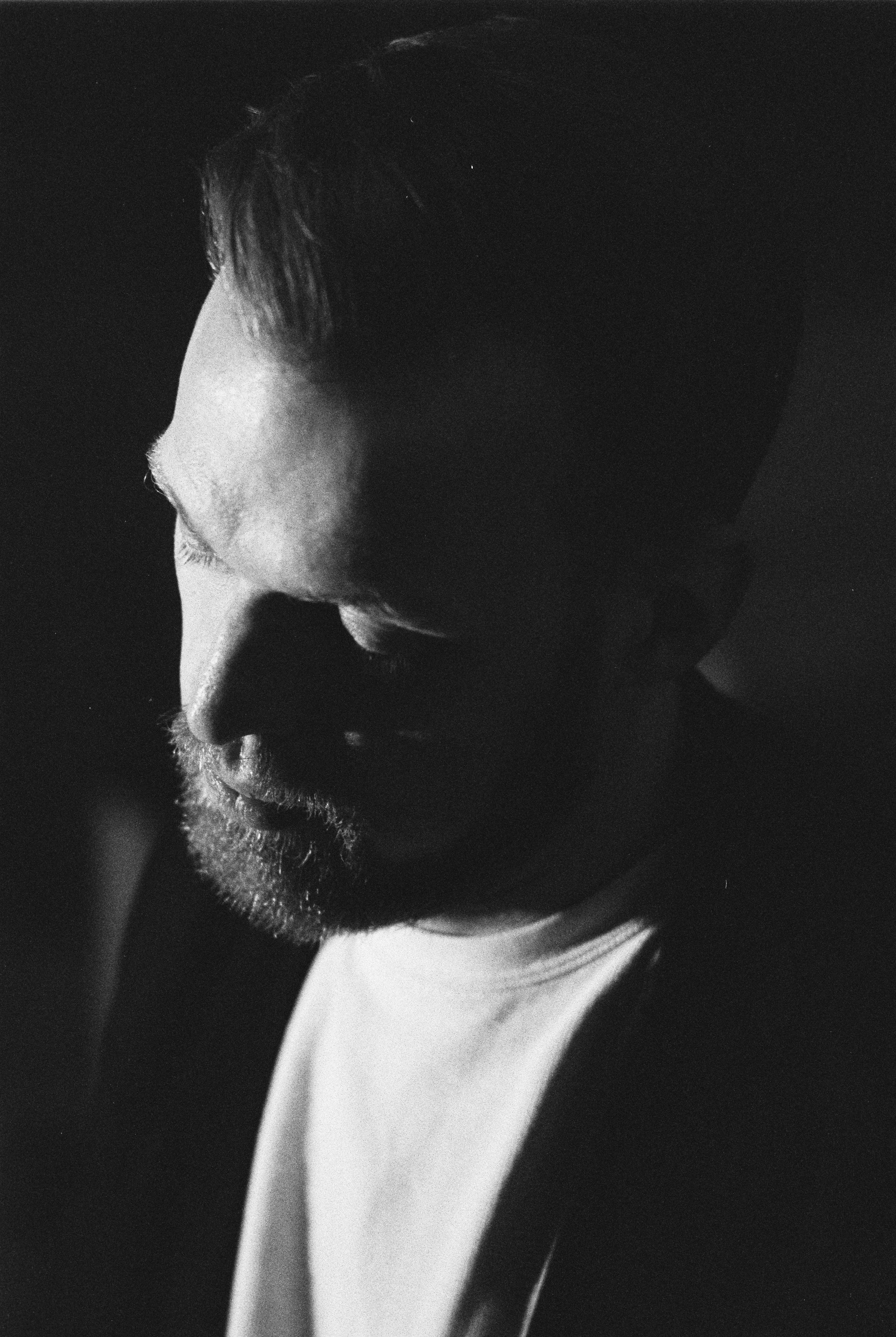 Schwarzweiß-Fotografie eines Mannes mit Glatze und Bart, nachdenklich mit Blick nach unten, halb im Schatten, dunklem Hintergrund.