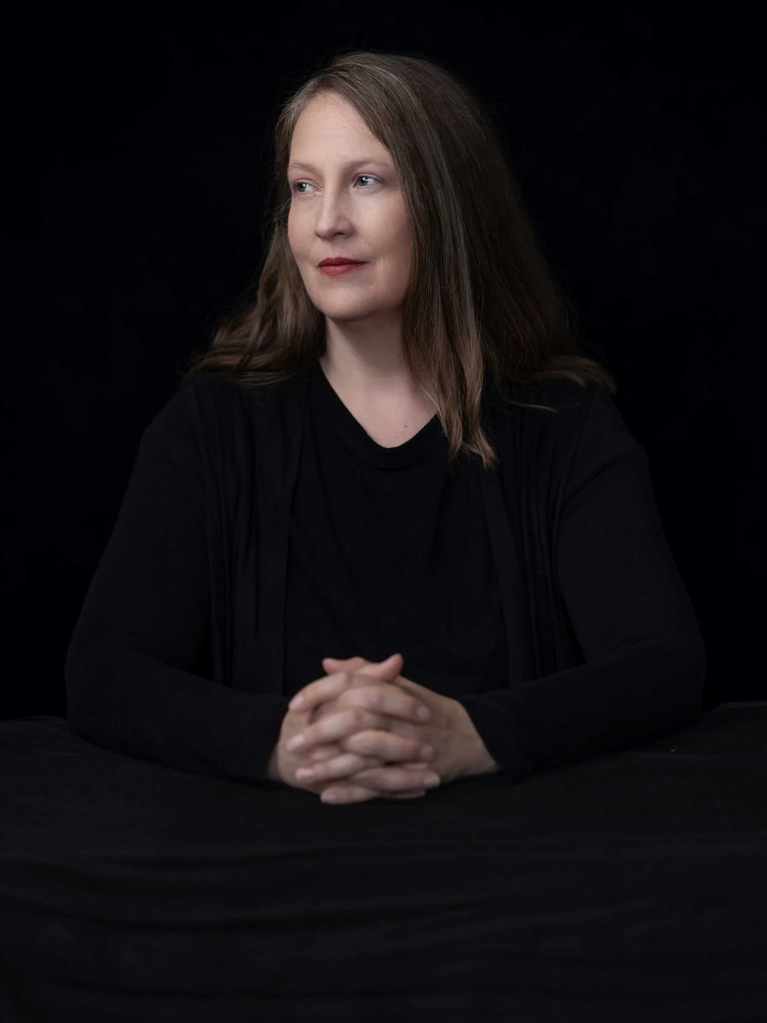 Author photo of JR Fenn looking to the side with hands clasped against a black background.