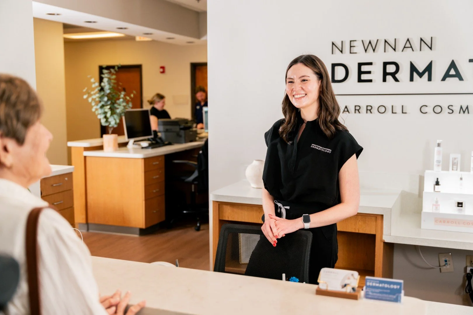 A woman wearing a black uniform smiling at a customer in a skincare clinic reception area.