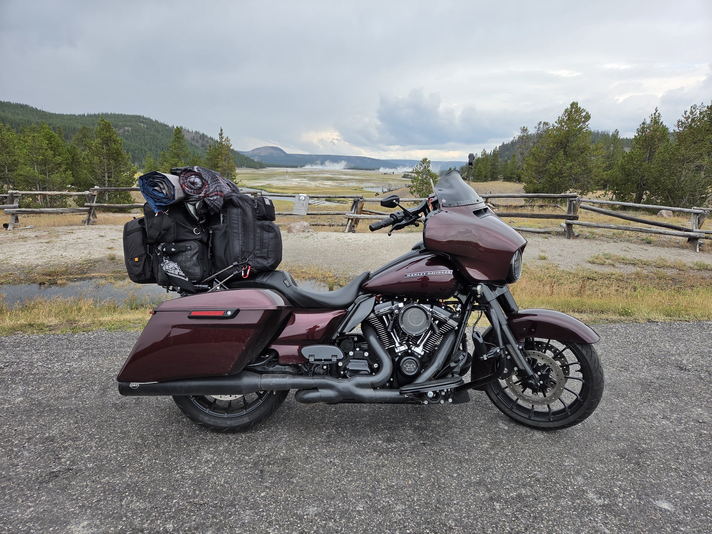 A maroon Harley Davidson motorcycle with luggage and blankets on the seat, parked on a gravel area overlooking a geothermal hot spring or geyser field with steam rising in the background, surrounded by forest and mountains under cloudy skies.