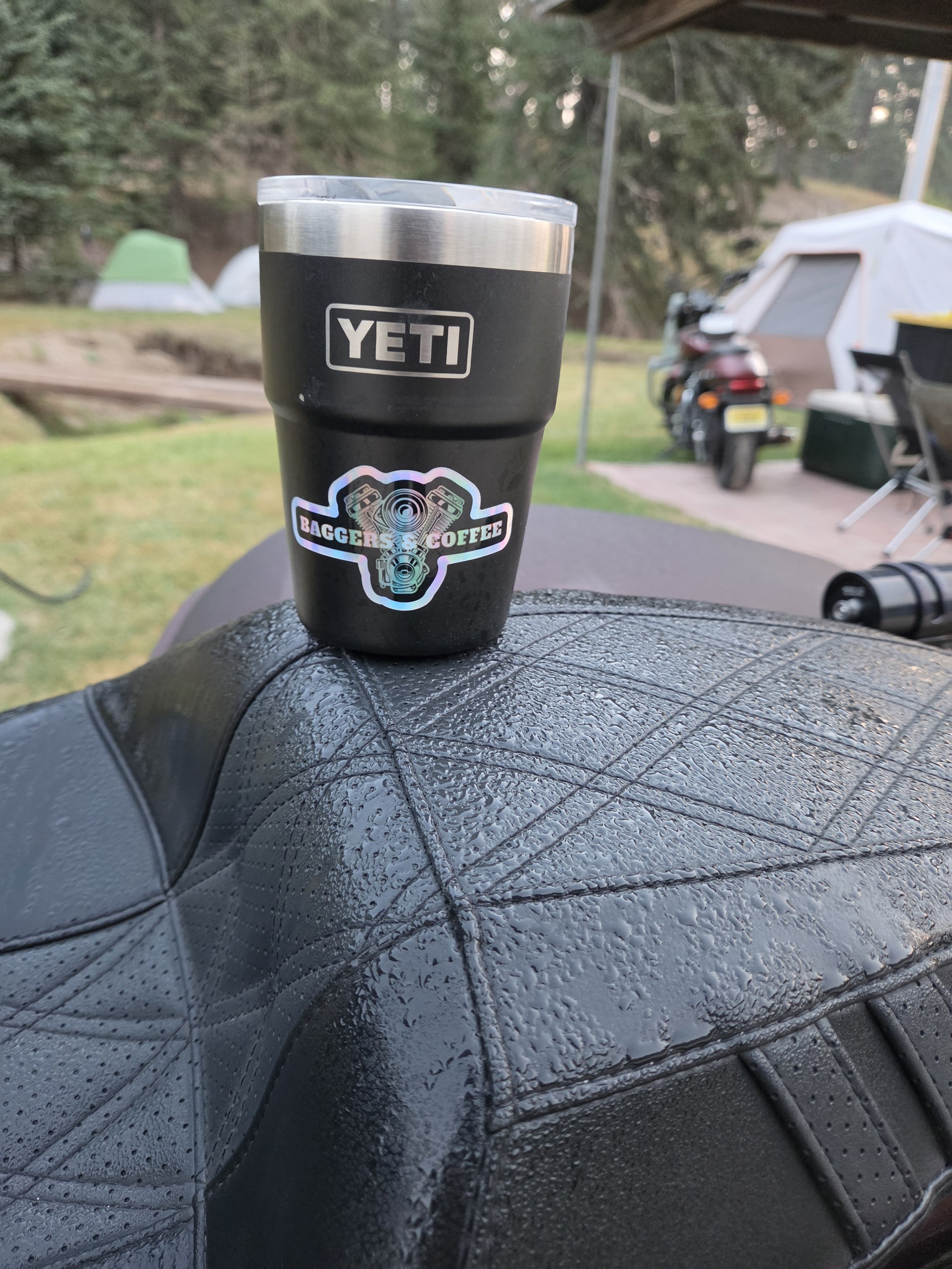 A black YETI travel mug with rain droplets on a wet black surface, with tents and trees in the background at a campsite.