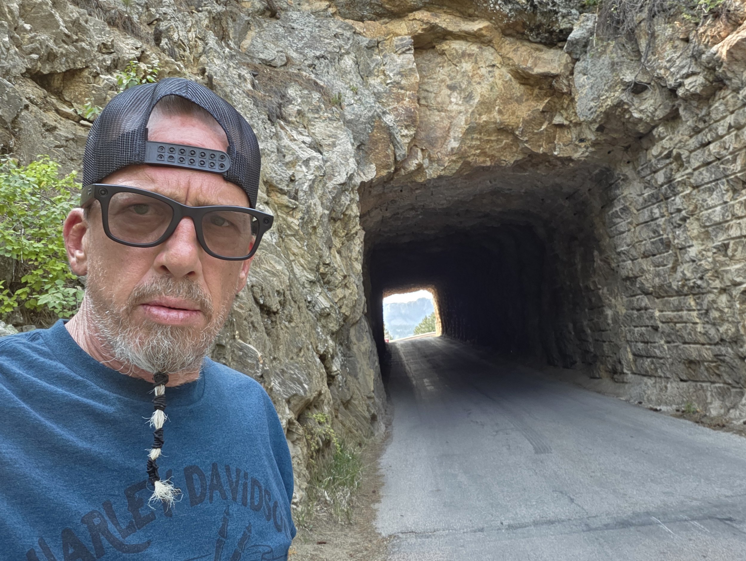 A man with glasses, a beard, and a backward baseball cap taking a selfie outdoors near a rocky tunnel.