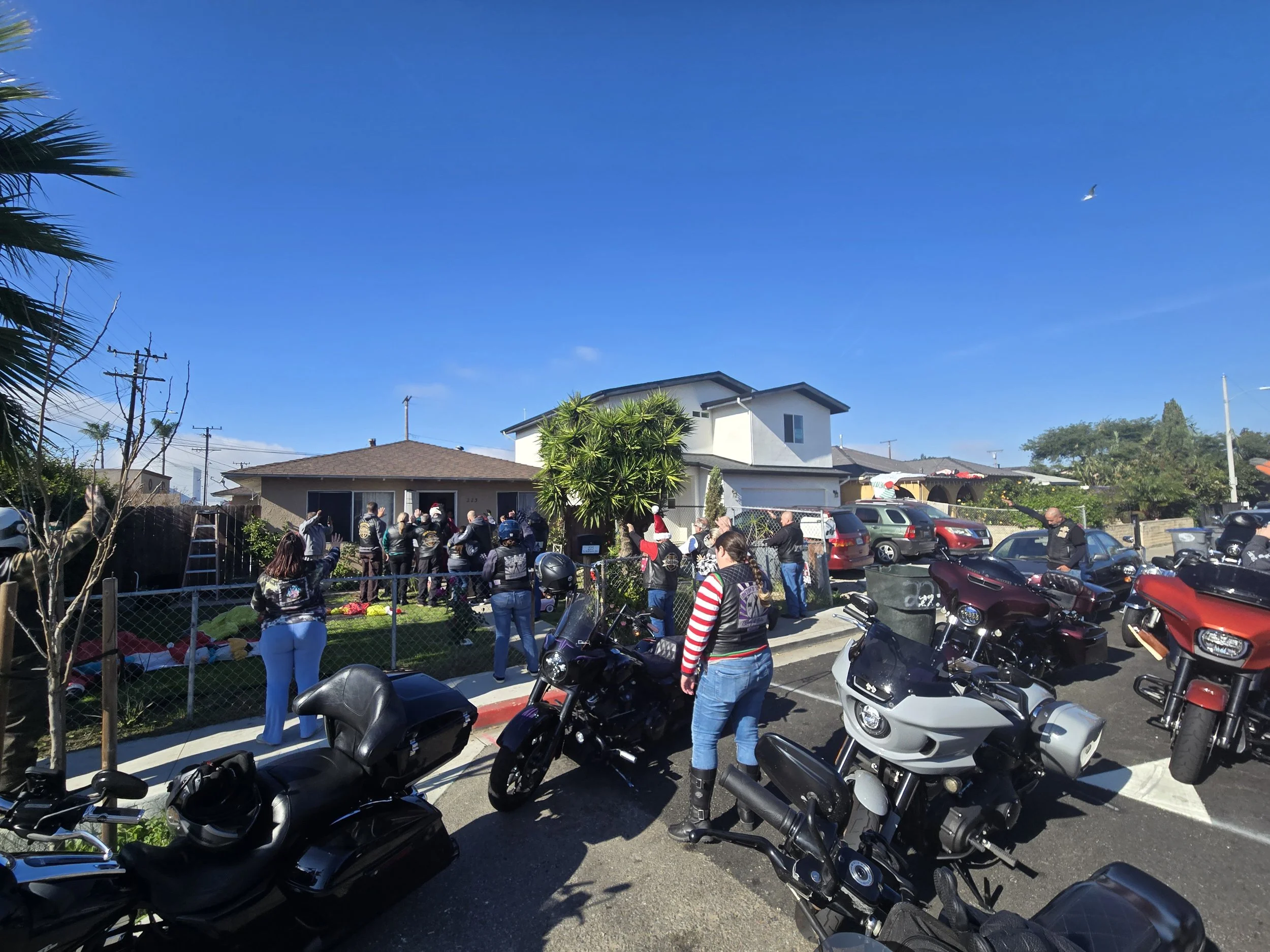 A group of people gather outside a house on a sunny day, some wearing Santa hats, with parking meters and motorcycles in the foreground, and a clear blue sky overhead.