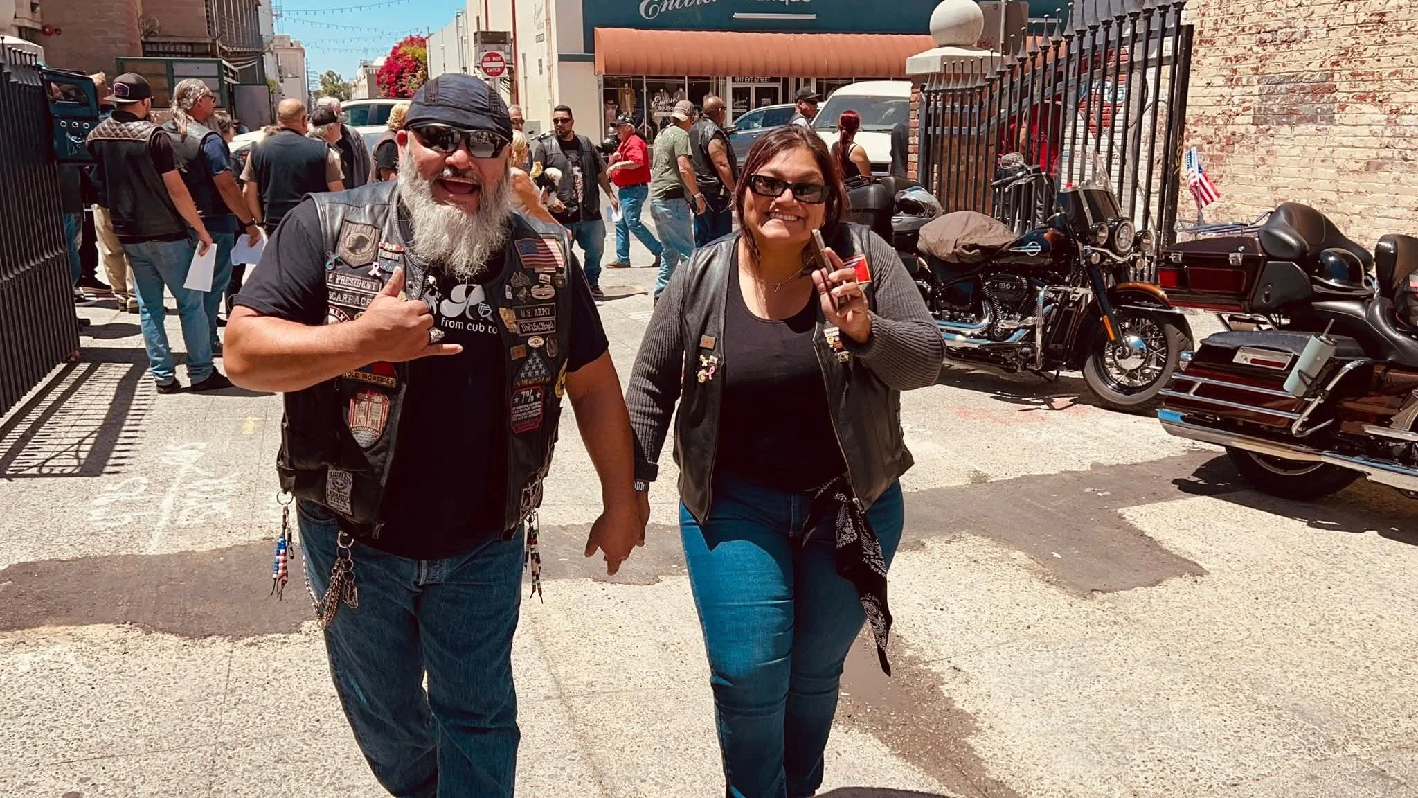 A man and woman standing on a city street, smiling and holding small objects, with a crowd and parked motorcycles behind them.