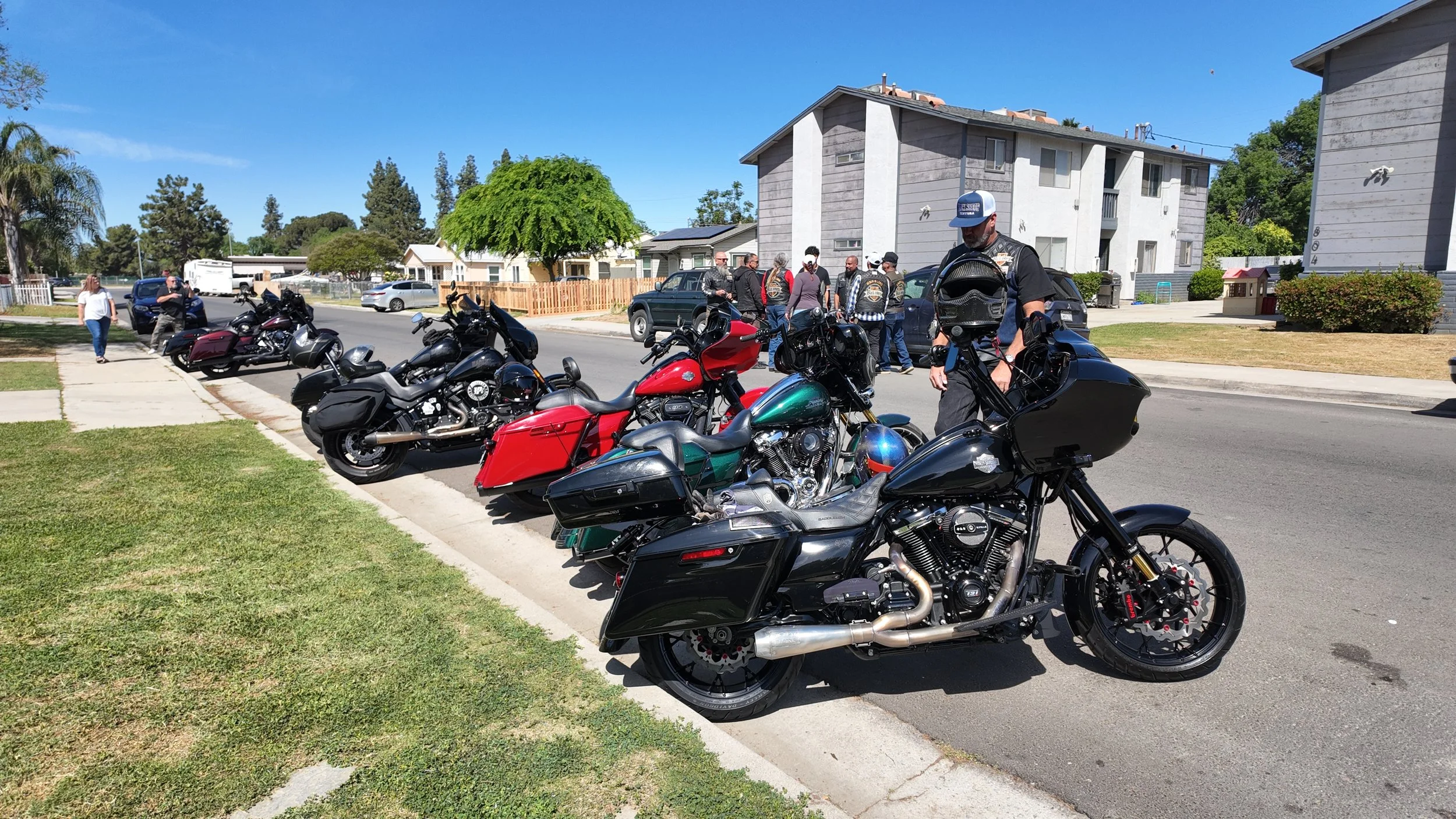 Row of parked motorcycles and a few cars on a residential street, with people standing and chatting in the background, under a clear blue sky.
