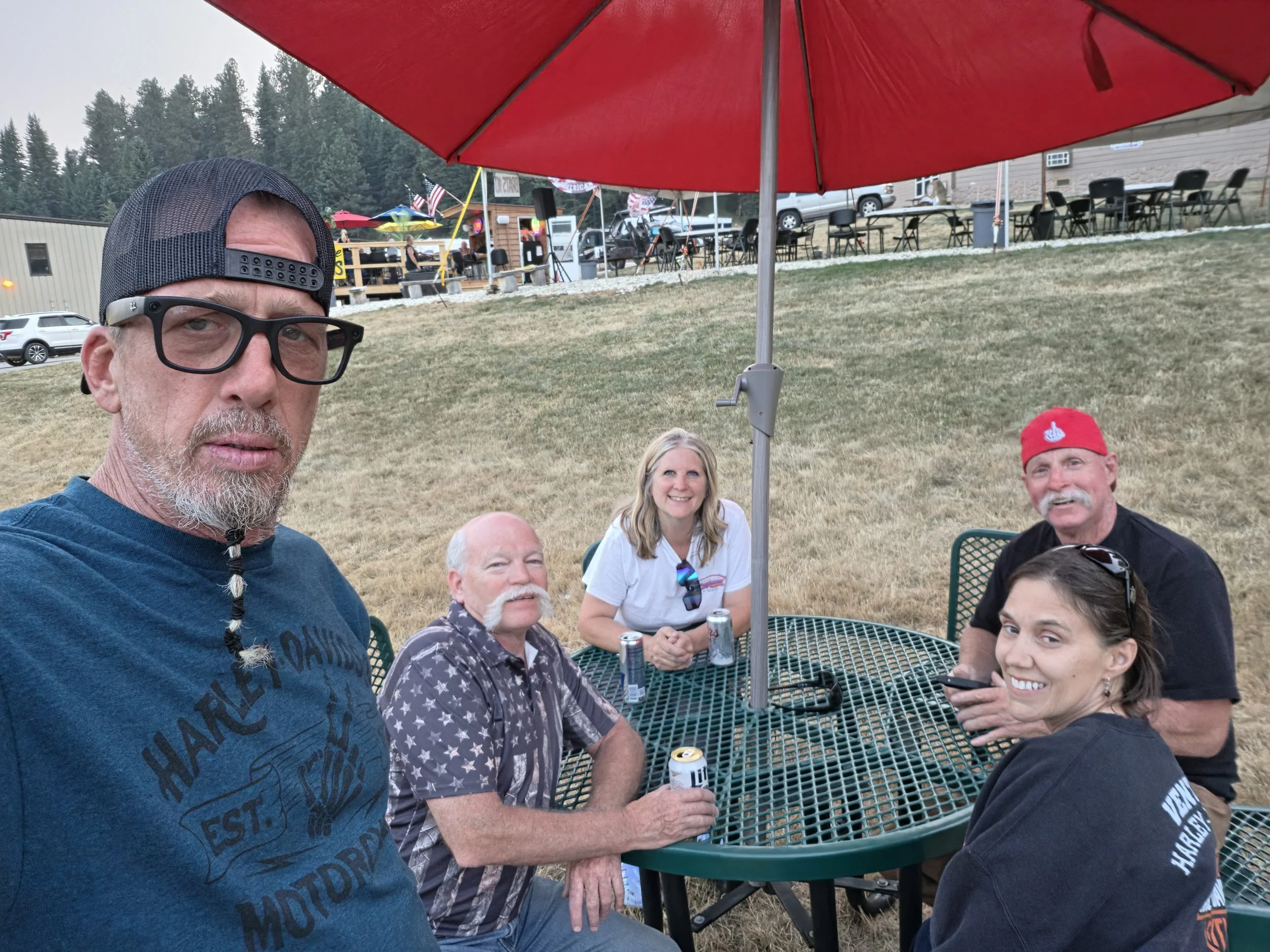 Group of people sitting at an outdoor table under a large red umbrella, smiling and holding drinks, with trees, vehicles, and festival tents in the background.