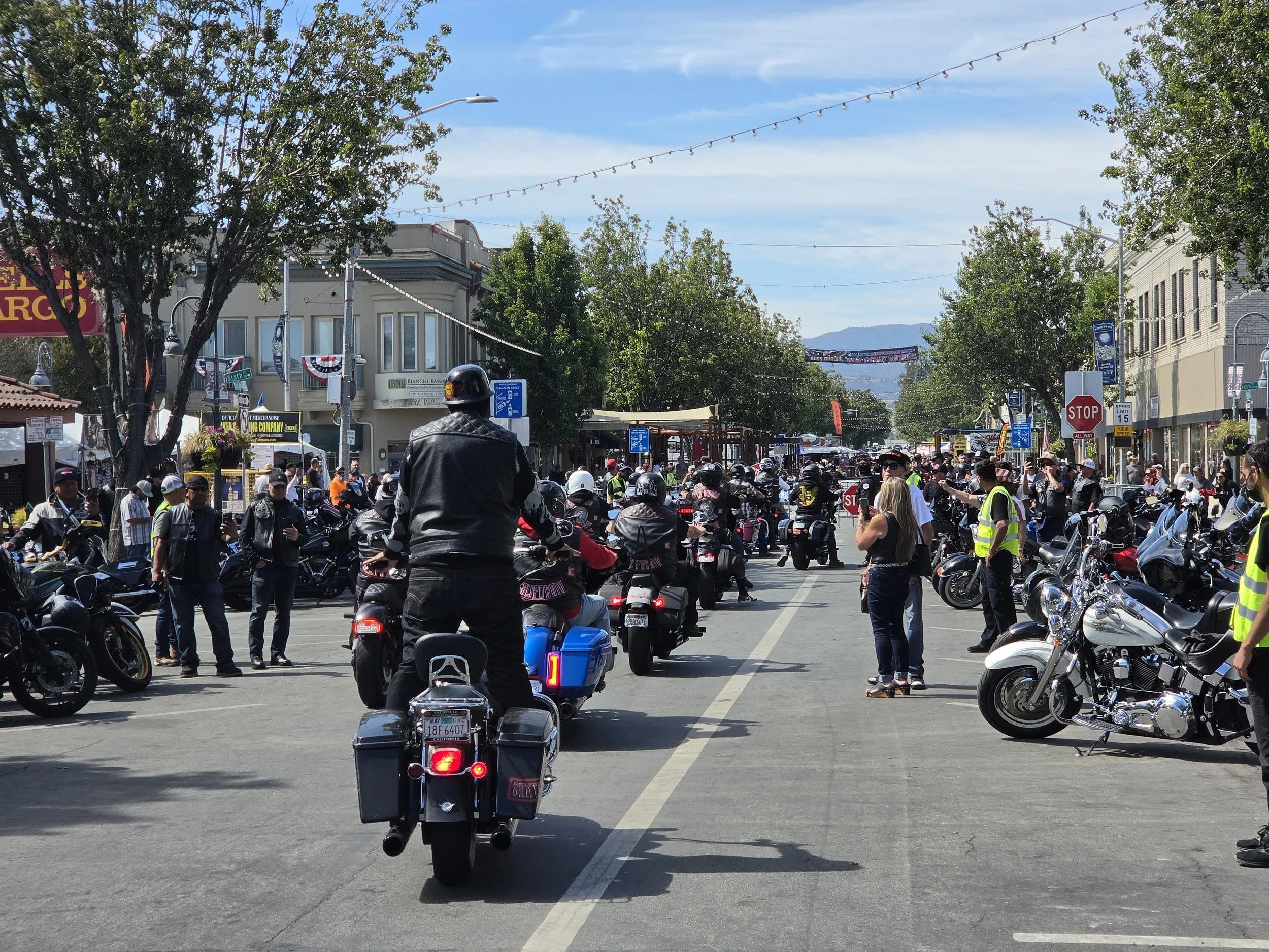 Street filled with parked motorcycles and scooters, with people standing around. Buildings, trees, and street signs are visible under a clear blue sky.
