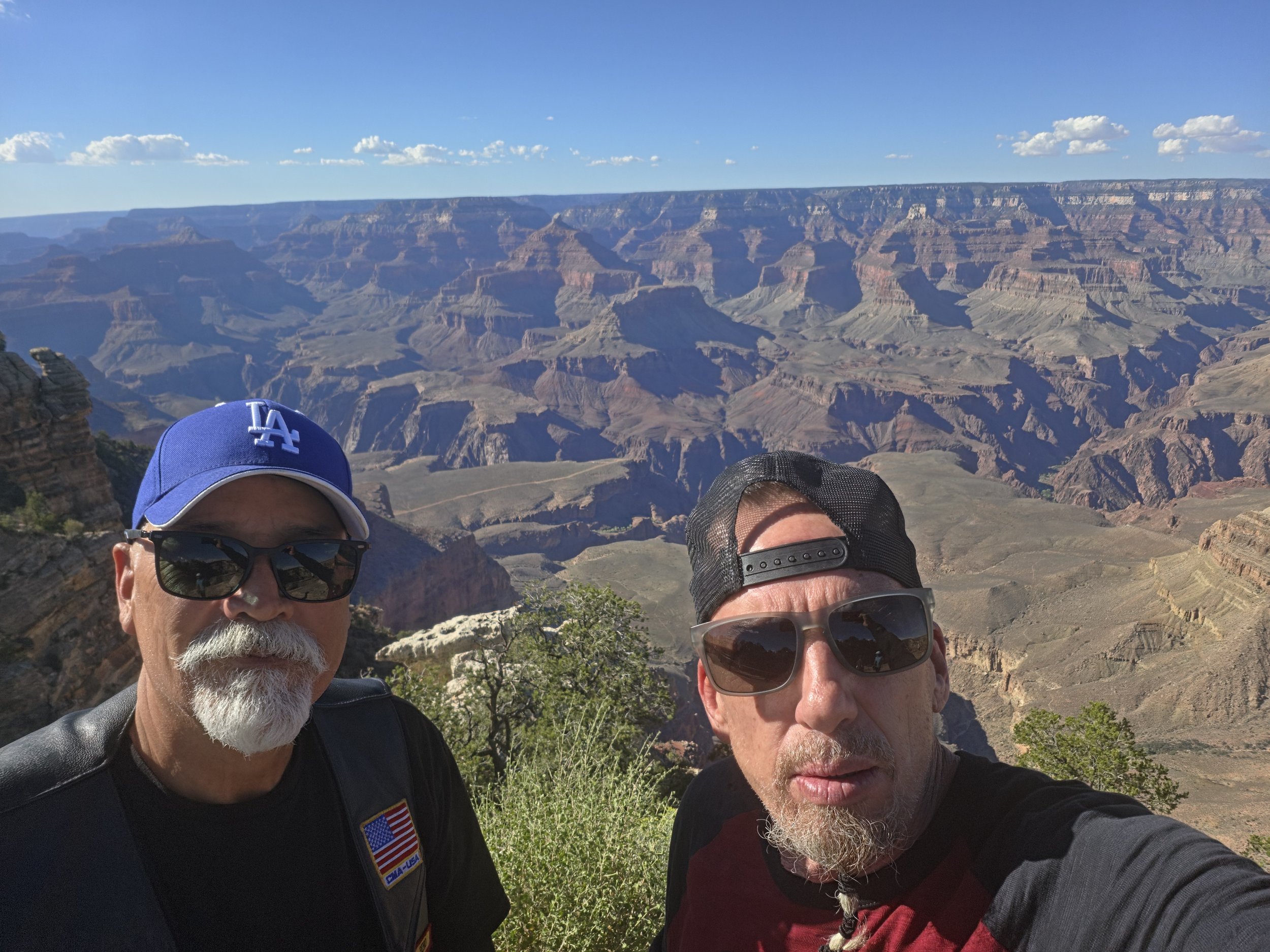 Two men taking a selfie at the Grand Canyon. The man on the left has a white beard, sunglasses, a blue cap, and a black jacket. The man on the right has a beard, large sunglasses, a black cap, and a red shirt. The background features the vast landsca