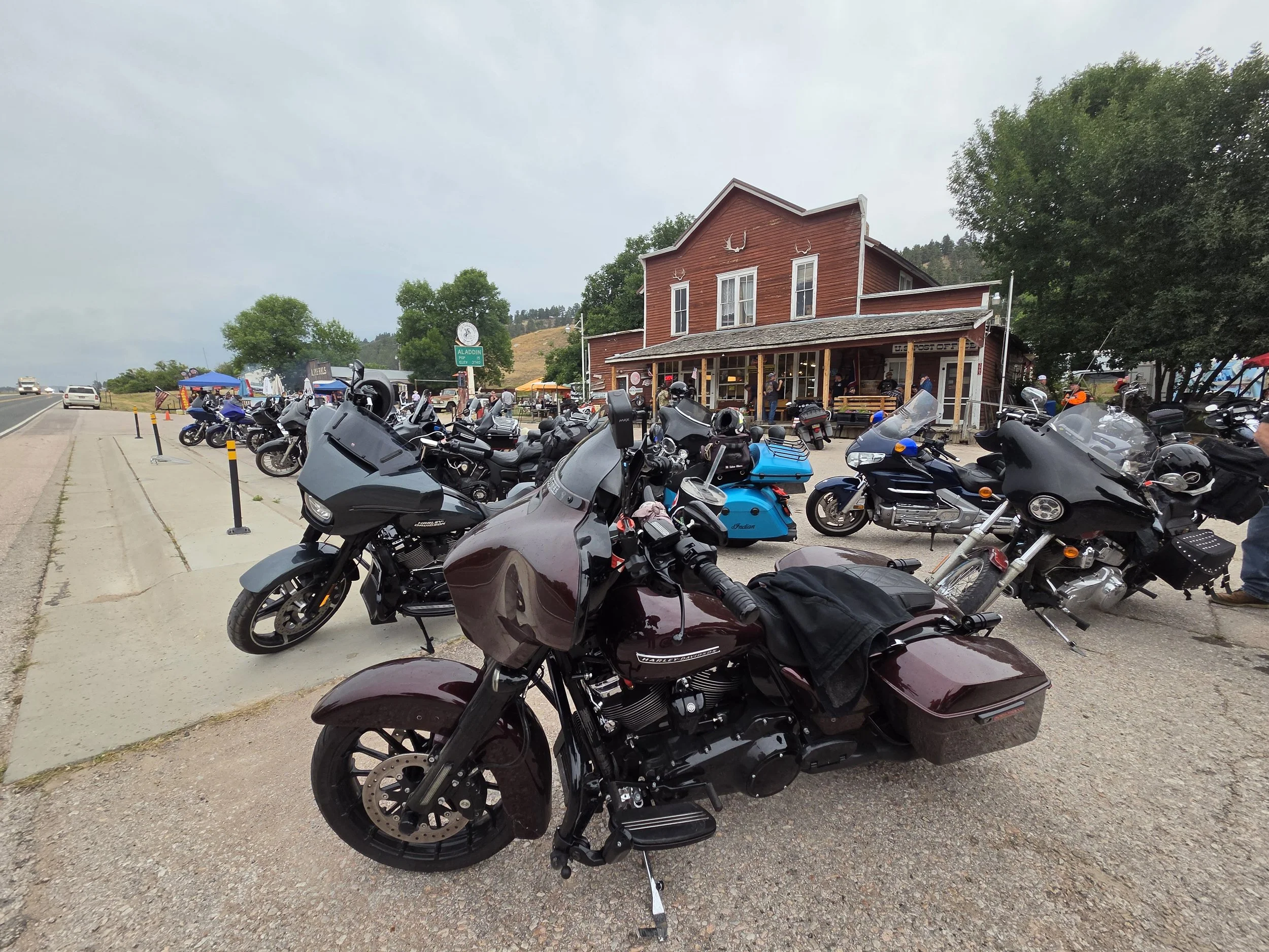 A parking lot filled with black motorcycles in front of a red wooden building with deer antler decorations on the facade. There are people sitting on the porch, and a sign indicating the location as Aladdin.