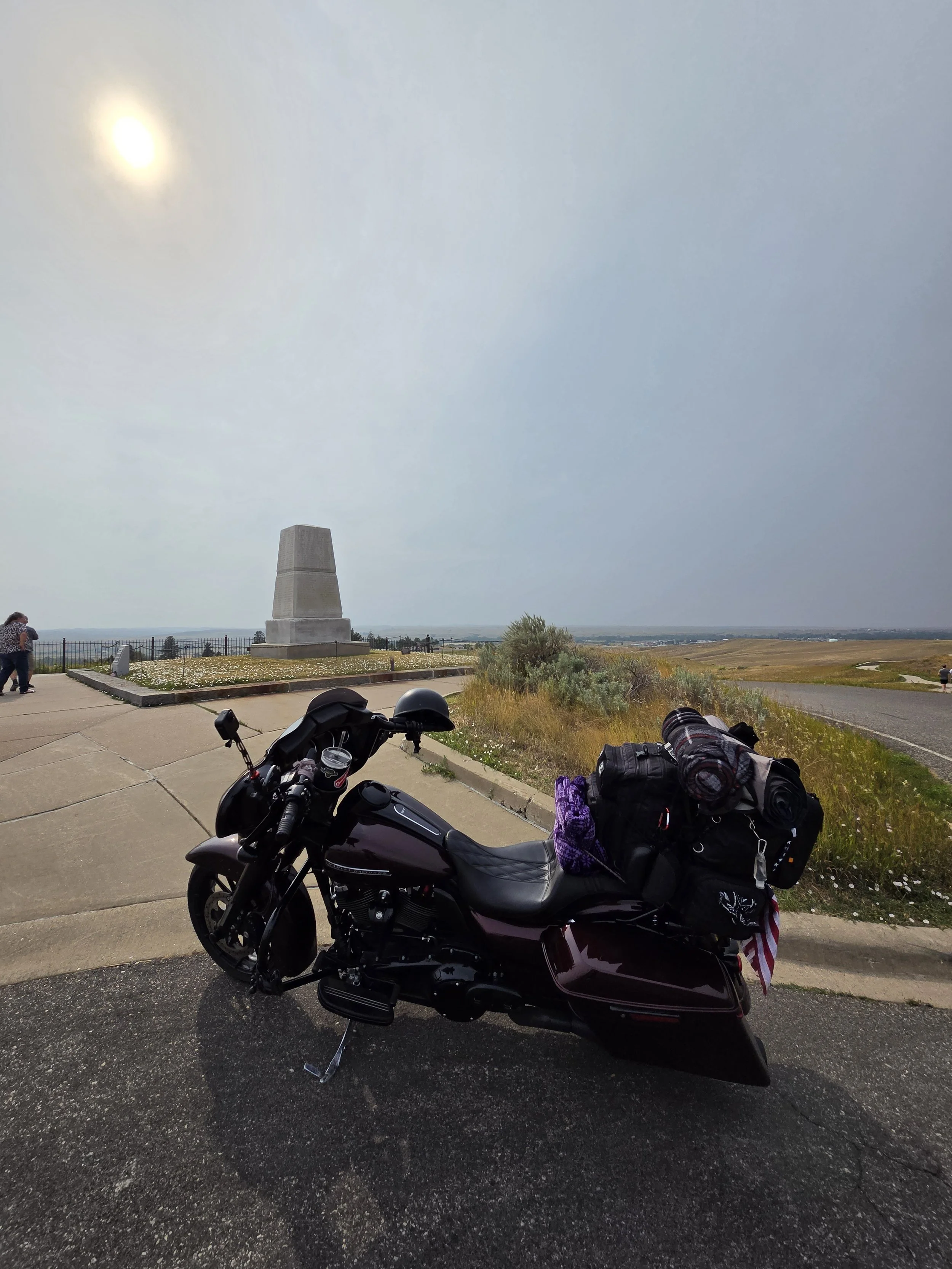 Motorcycle parked on the side of the road with luggage and gear attached, overlooking a scenic landscape with a monument, open fields, and a partly cloudy sky.