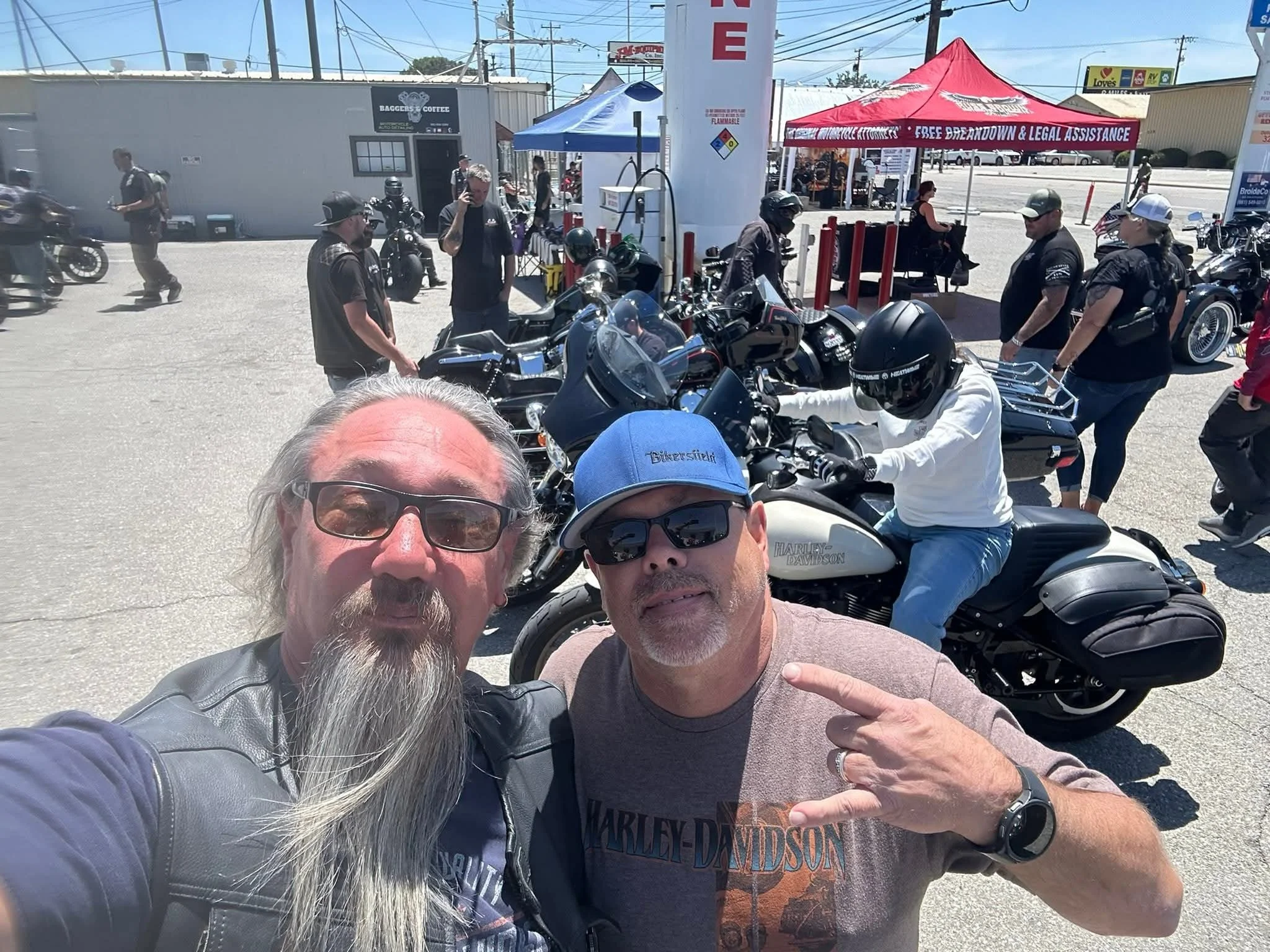 Two men taking a selfie in front of motorcycles at an outdoor event, with tents and people in the background.