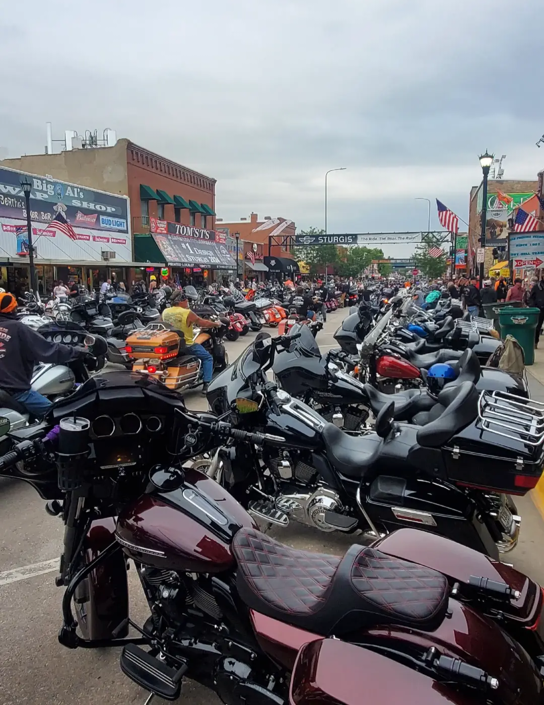 Crowded street filled with motorcycles parked on both sides and people walking along the sidewalk. Shops and signs visible, including a bar, shop, and various flags hanging across the street. Overcast sky.