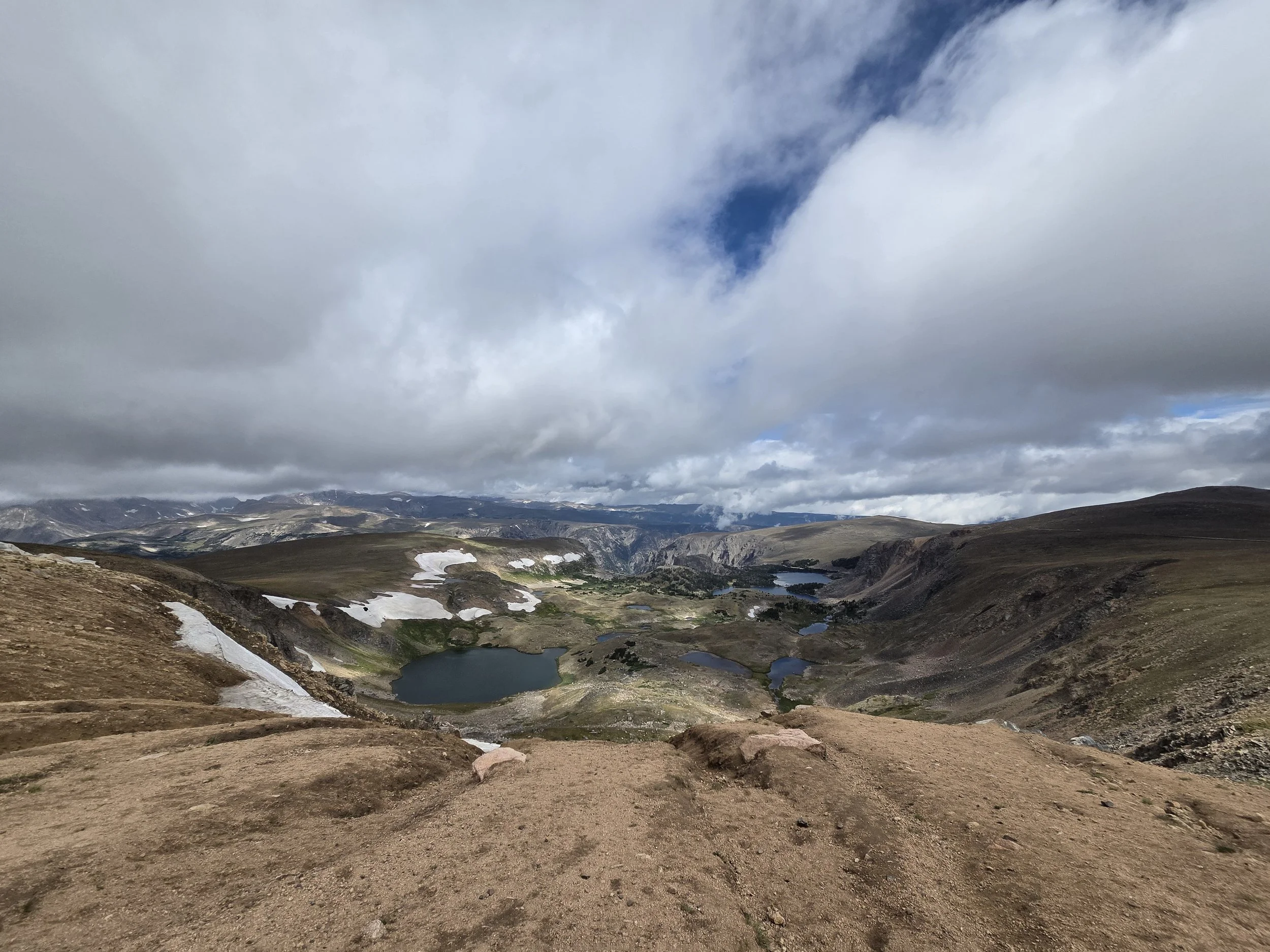 A mountainous landscape with several lakes, patches of snow, rocky terrain, and a cloudy sky overhead.