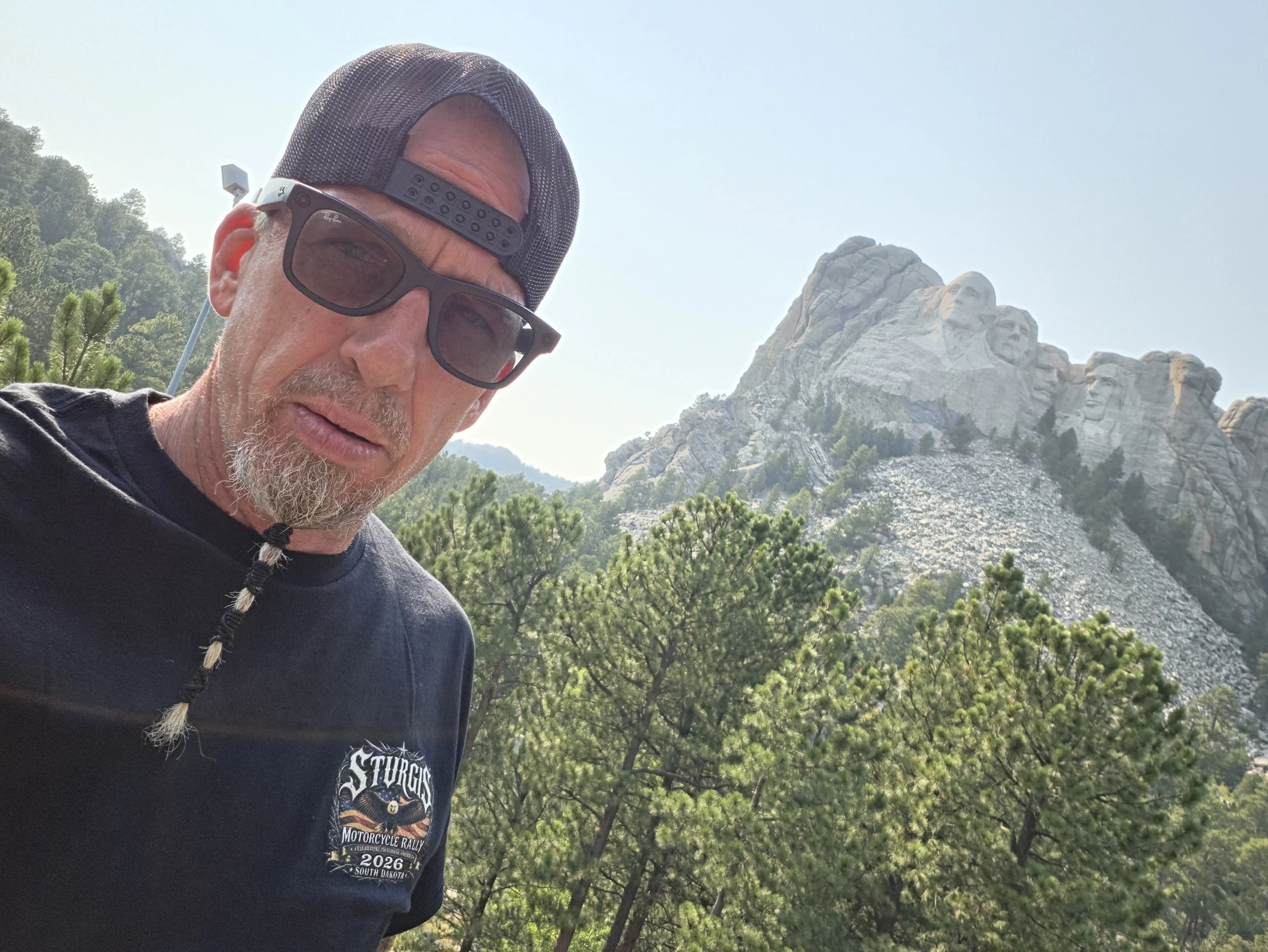 A man taking a selfie outdoors with Mount Rushmore in the background, partly obscured by trees.