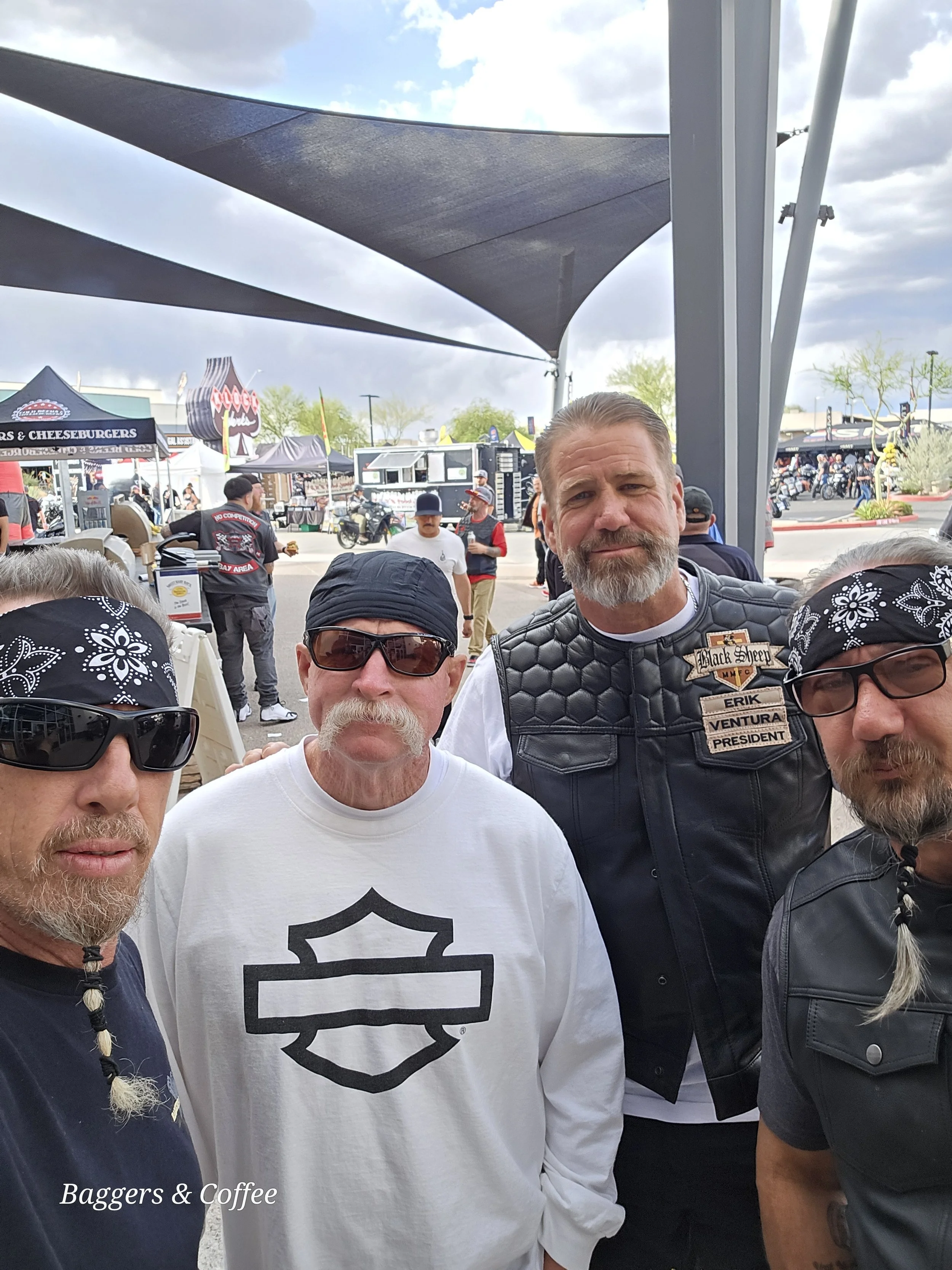 A group of men gathered outdoors at a motorcycle event or gathering with tents and motorcycles in the background. One person wears a black leather vest with a name tag, and another wears sunglasses. The sky is partly cloudy.
