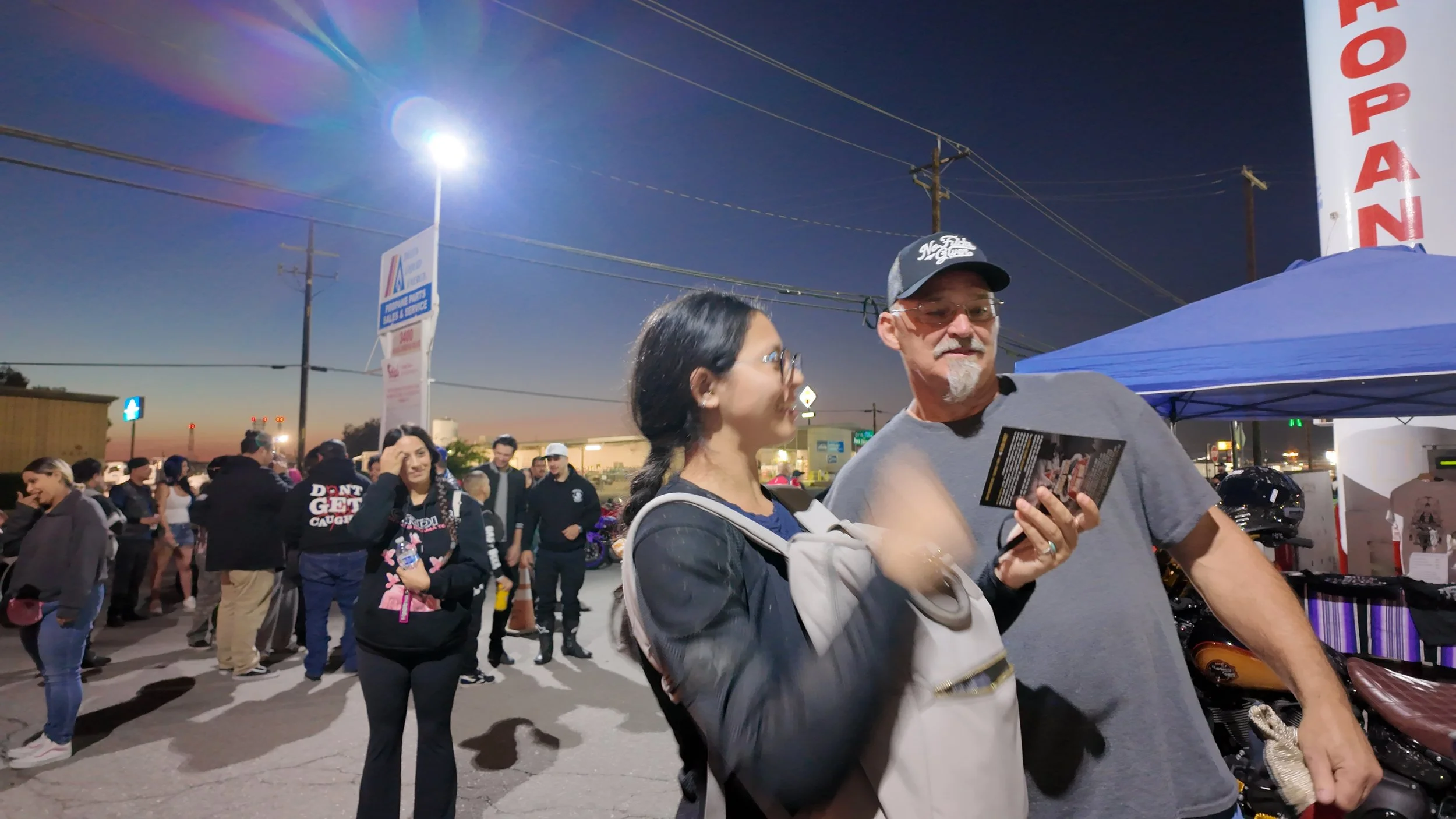 People gathered outdoors during evening, some chatting near a blue tent, with utility poles, sky, and signage in the background.