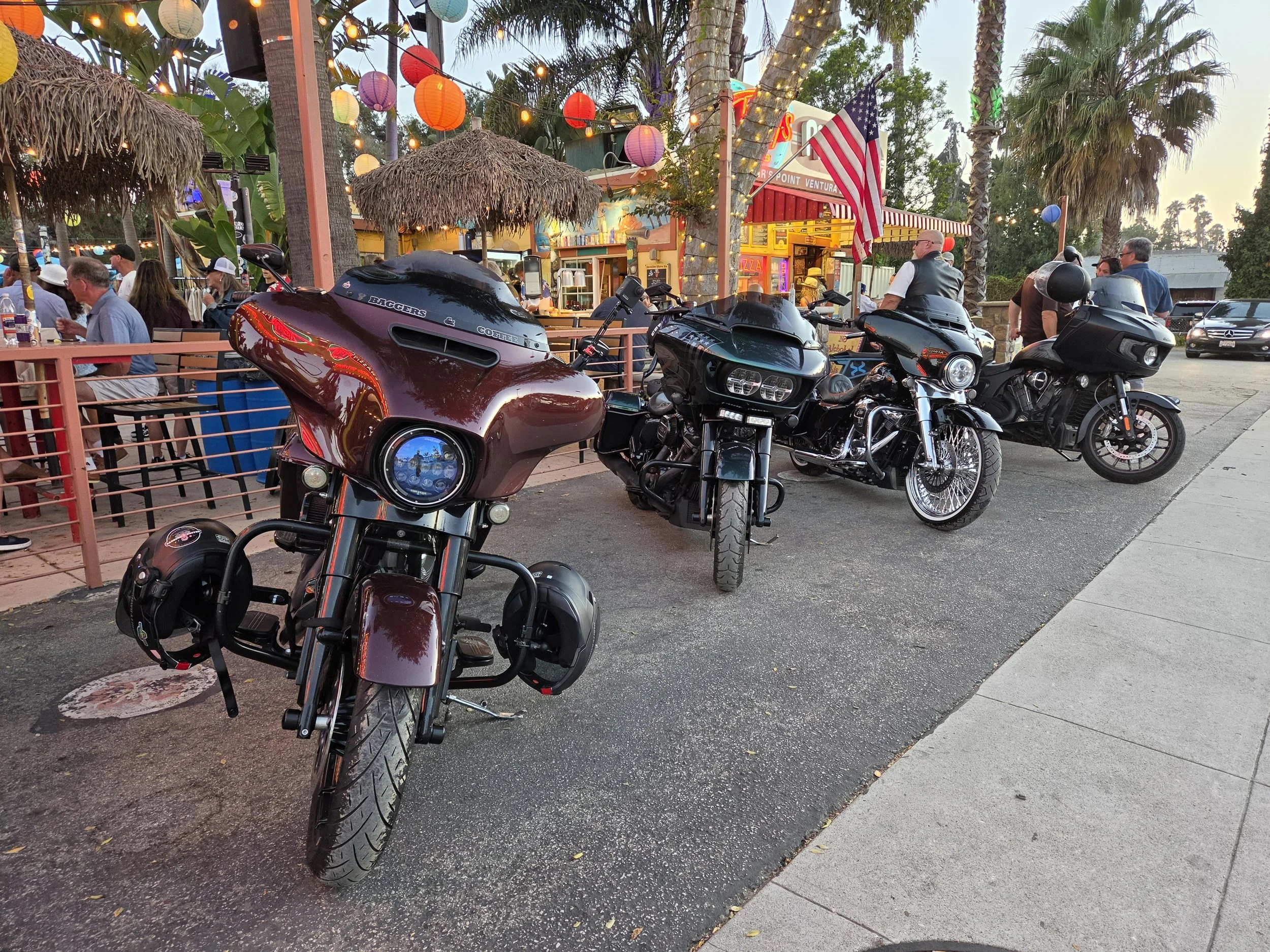 Parking lot with three black motorcycles in the foreground, a restaurant with colorful lights and paper lanterns hanging from trees in the background, and several people sitting and standing around, some near the motorcycles