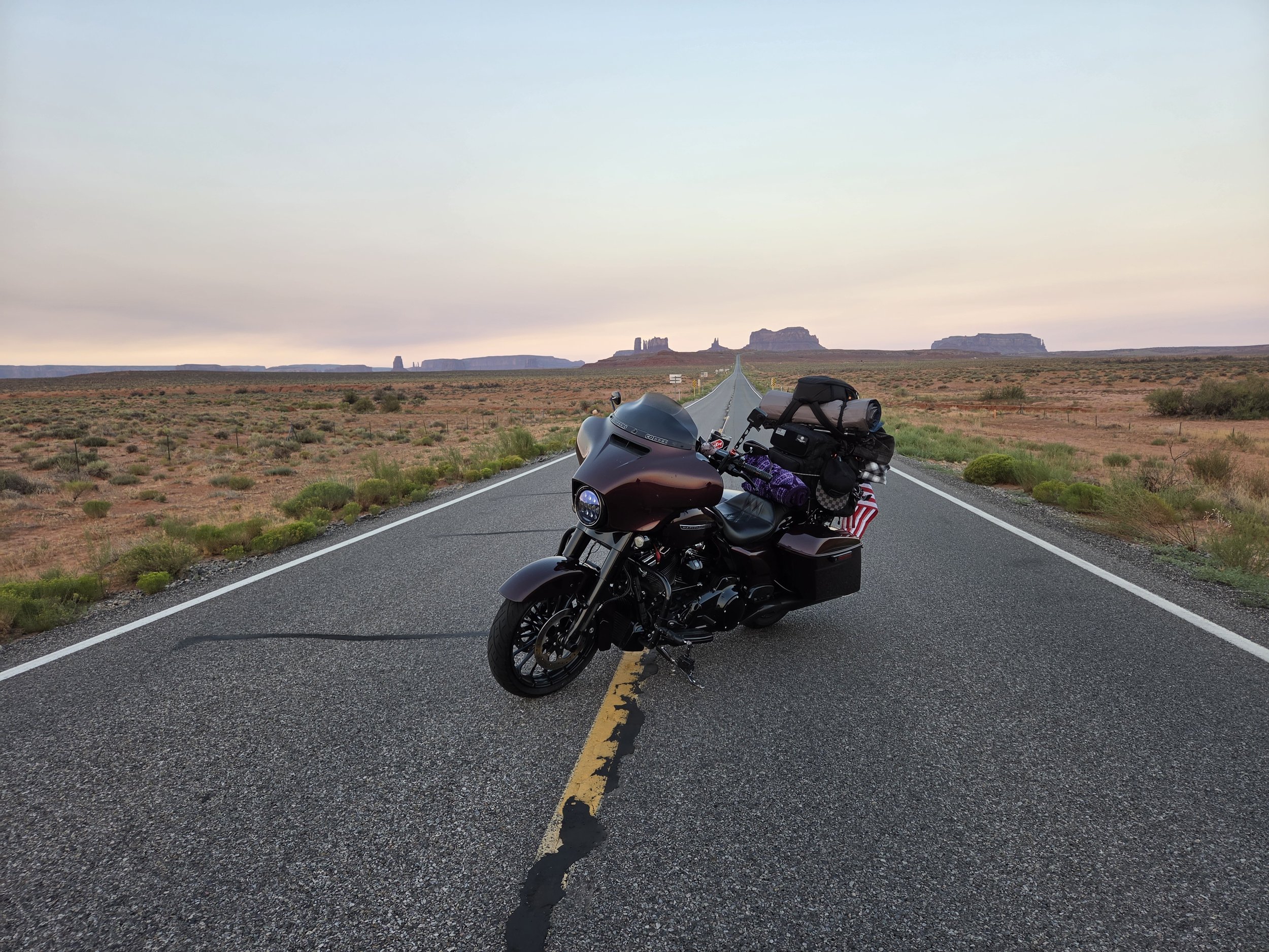 A black motorcycle parked on a long, straight desert road with a cracked yellow line running down the center. The landscape features sparse vegetation and distant mesas under a pastel-colored sky.