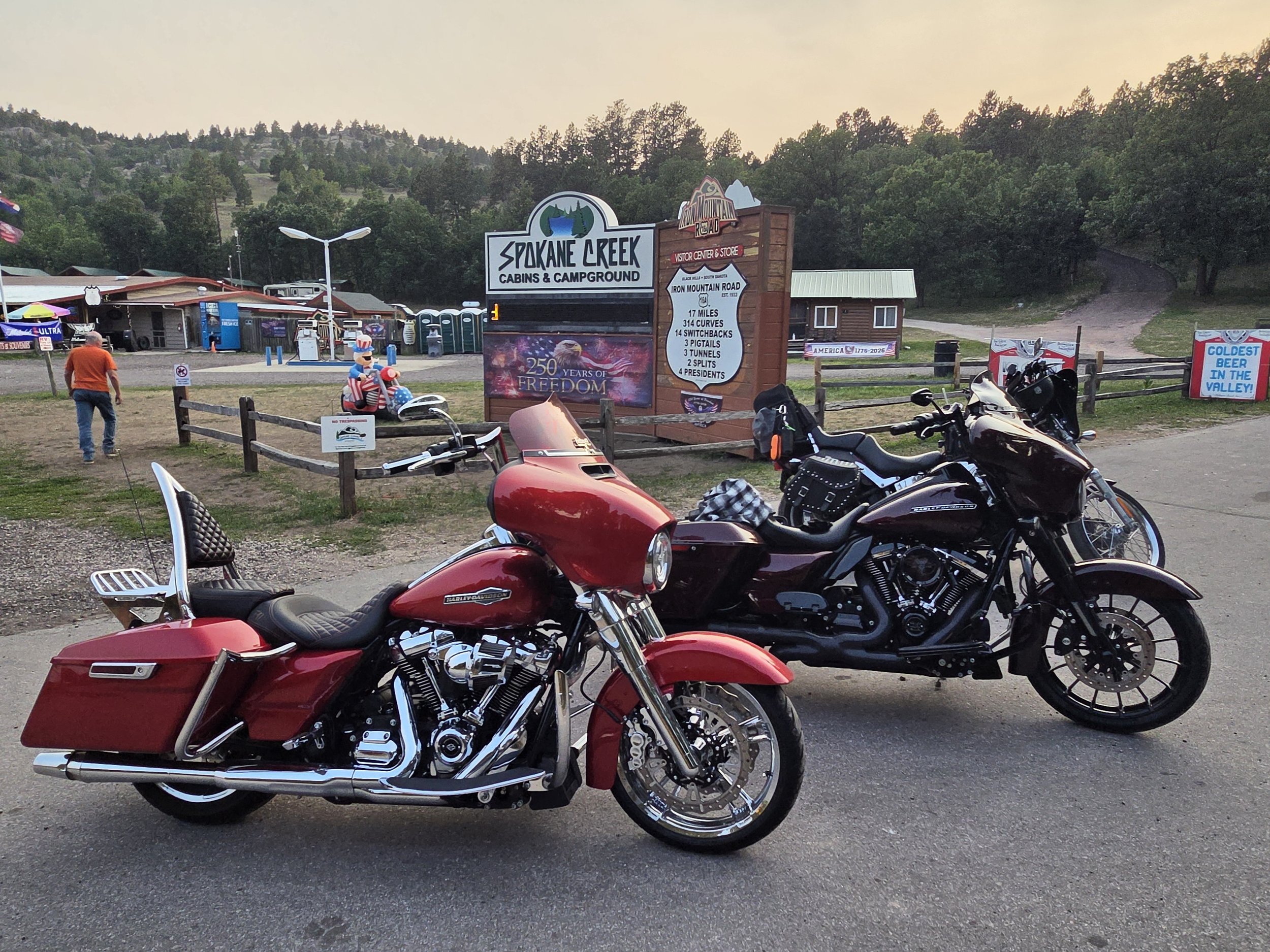 A parking lot with several motorcycles in the foreground, including a red Harley Davidson and a black motorcycle. In the background, there are signs for Spokane Creek Cabins & Campground, a small building, a few people walking, and trees on a hillsid