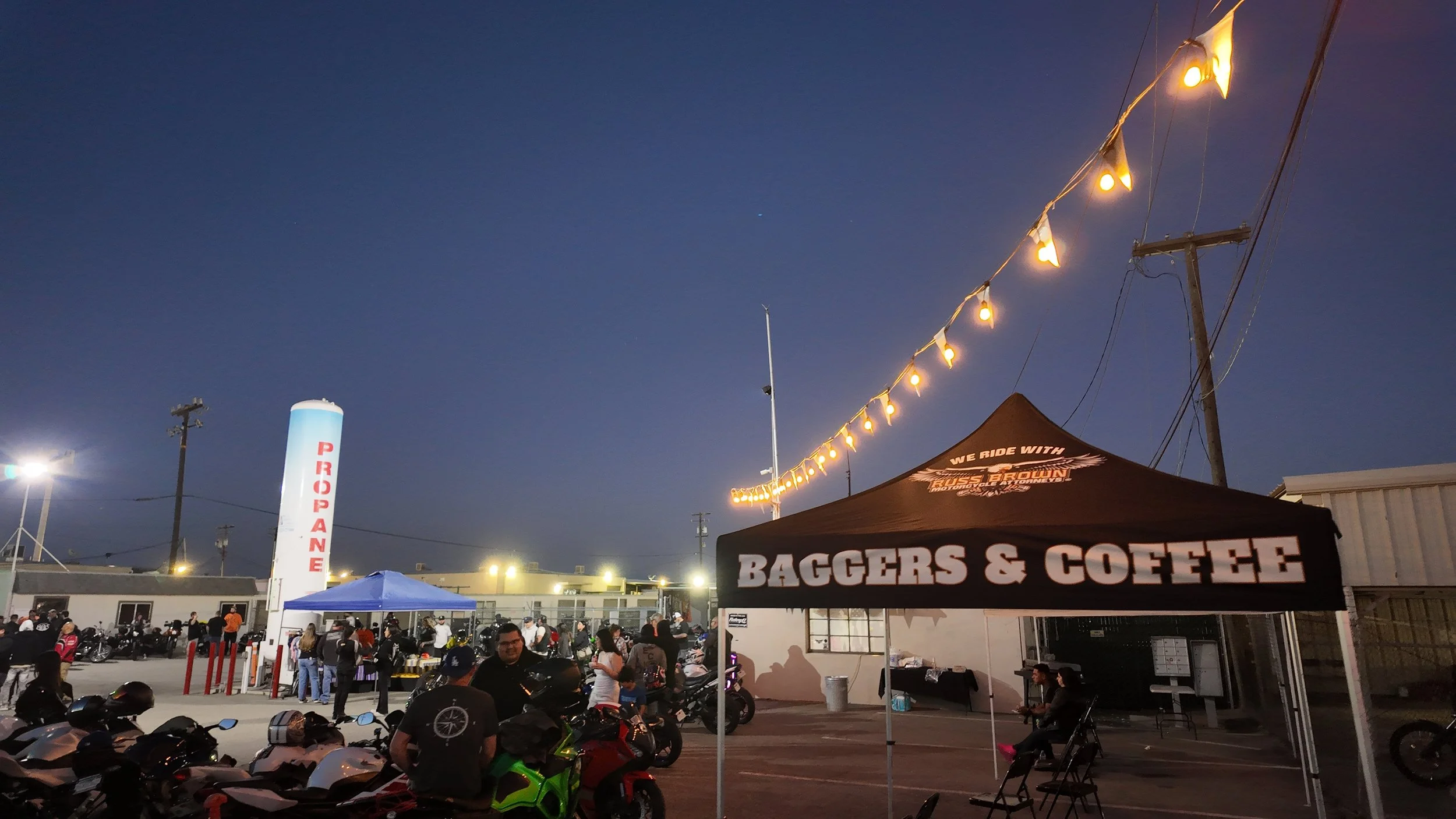 An outdoor event with motorcycles parked on a lot and a crowd of people. There is a tent with the words 'BAGGERS & COFFEE' and string lights hanging overhead. The background includes a large white column with red lettering, electrical poles, and a da