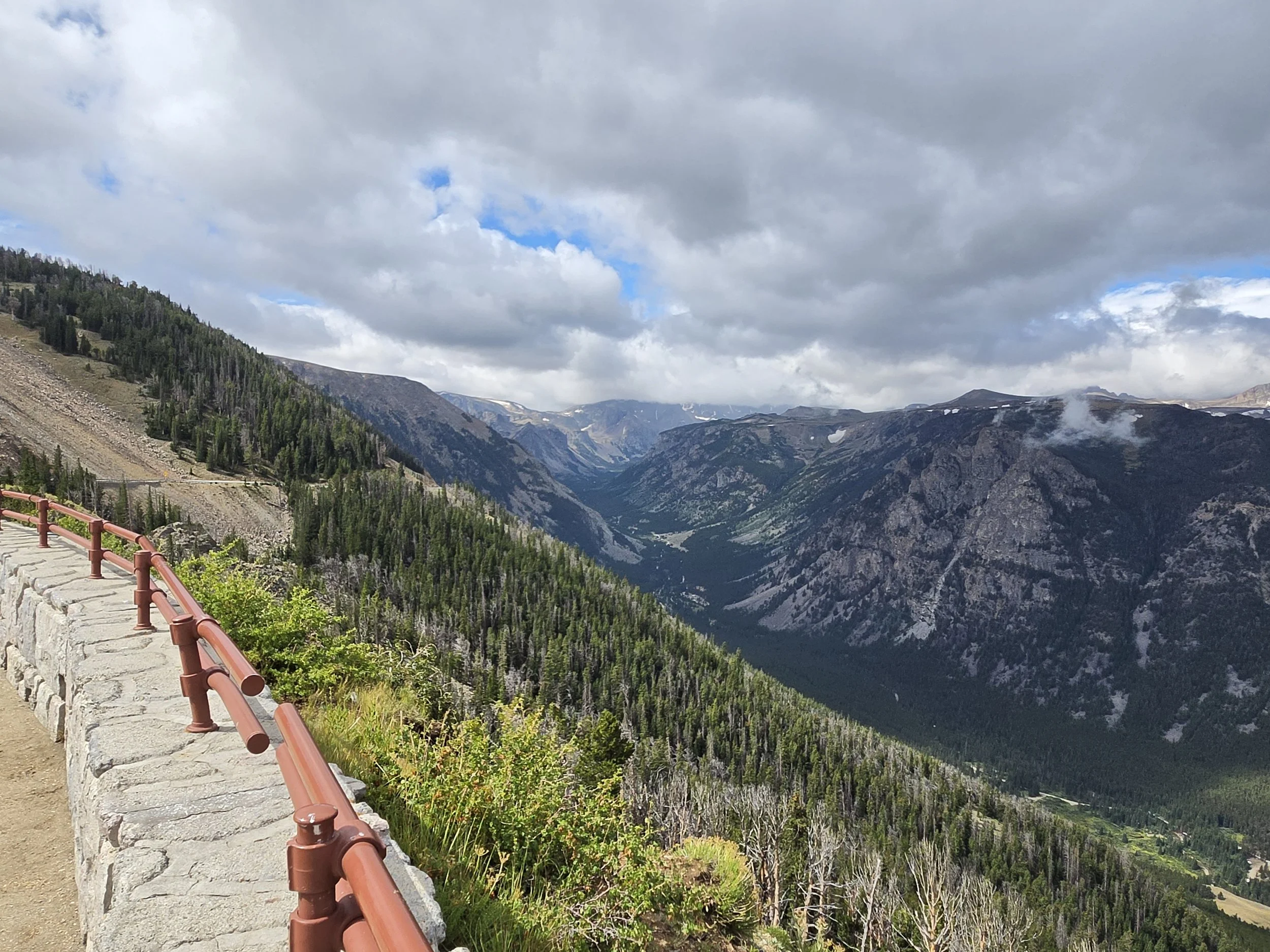 Scenic view of a mountain valley with clouds overhead, a winding river, and a scenic overlook with a stone and metal railing.