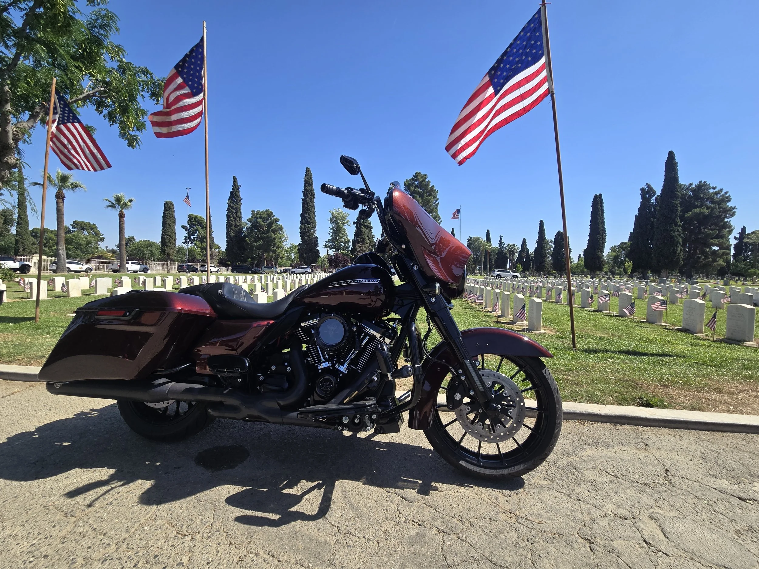 A black and red motorcycle parked in front of a row of small American flags at a cemetery, with tall trees and flags flying in the background.