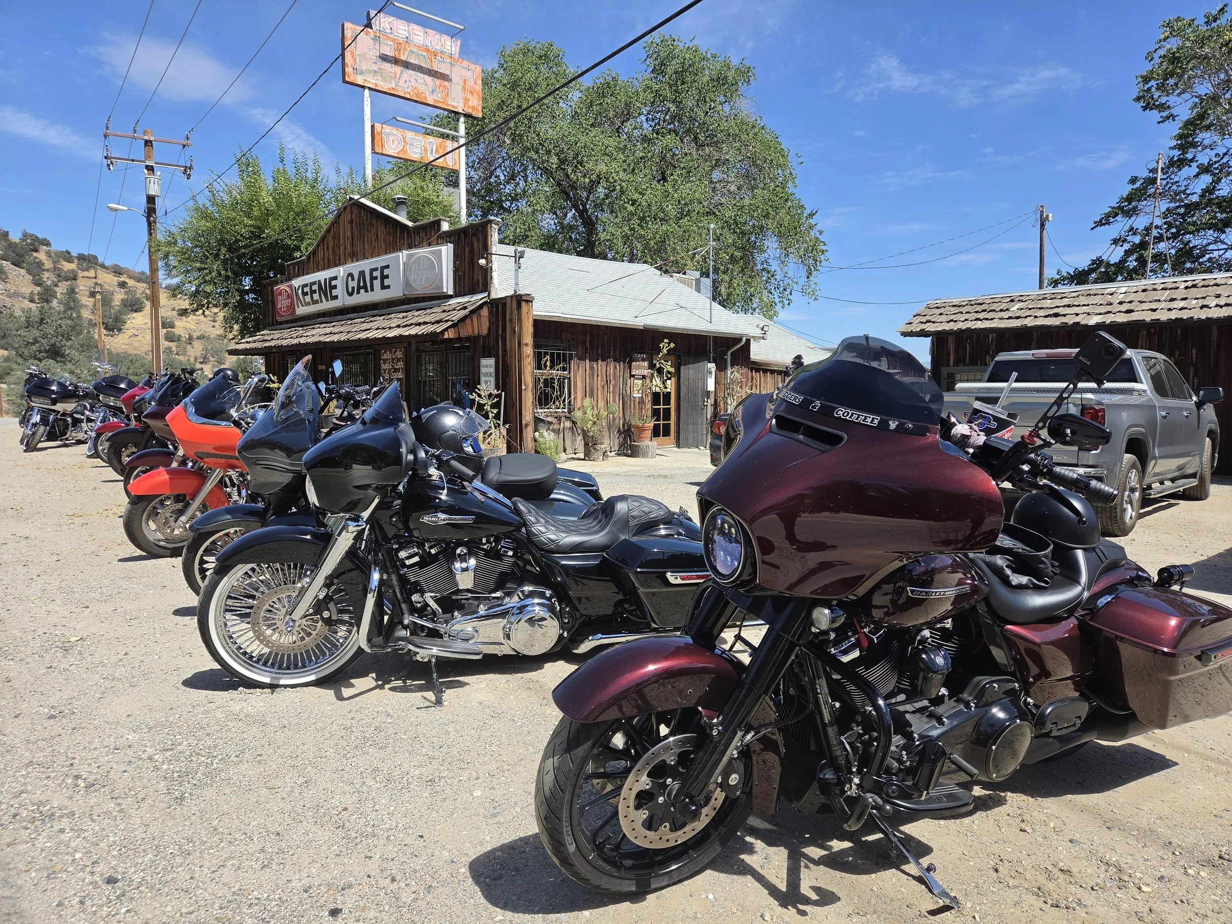 Vintage wooden building with a sign that reads 'Keene Cafe' and a rusted sign above it. In front, there are multiple parked motorcycles and a truck. The background features trees and a blue sky.