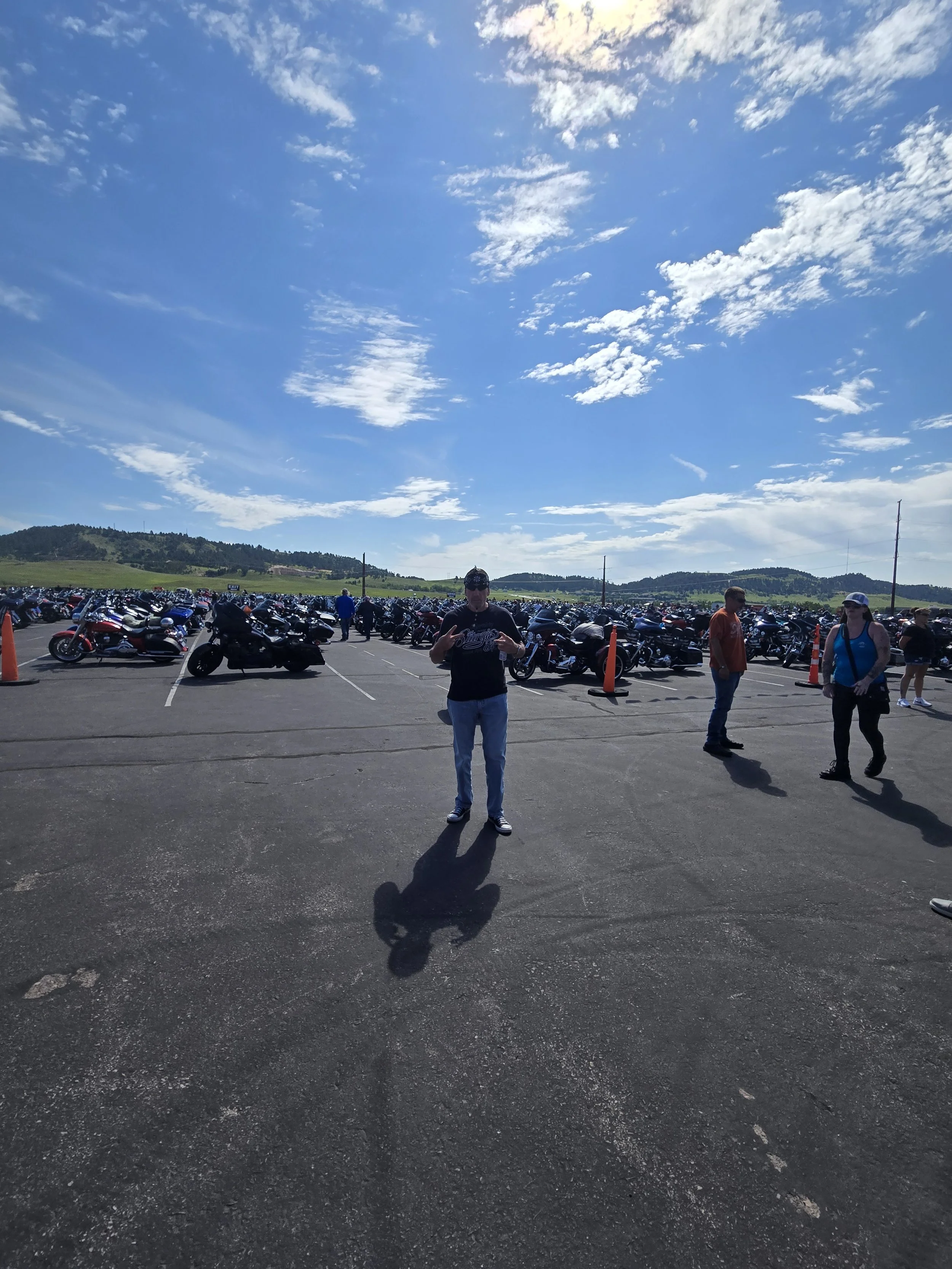Wide shot of a parking lot filled with motorcycles, with several people walking among the bikes under a partly cloudy sky.
