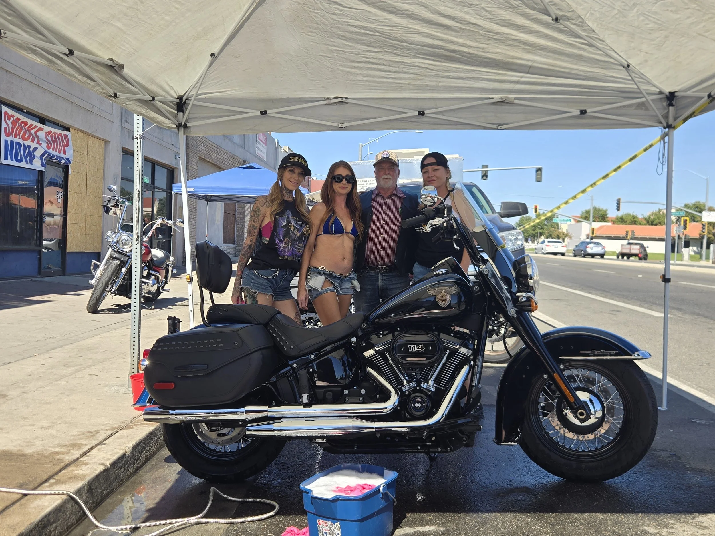 Group of four people standing behind a parked black motorcycle under a white canopy at an outdoor event. One woman is in a revealing outfit, and others are dressed casually. There are other motorcycles and tents nearby, with buildings and traffic lig