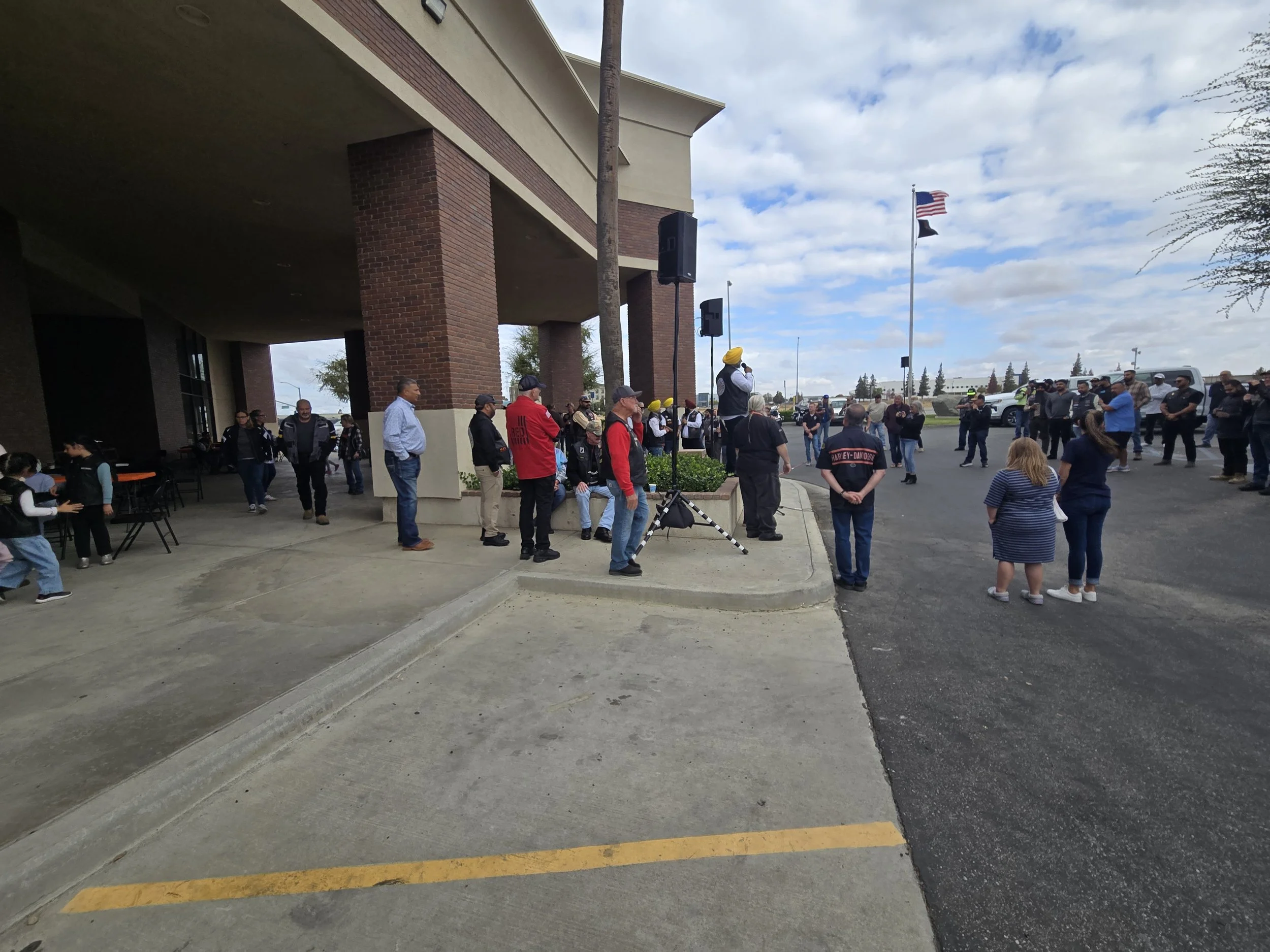 A crowd of people gathered outside a building with brick pillars and an overhang, some volunteer or event staff wearing uniforms, one person speaking into a microphone with a yellow turban, while others stand or sit nearby. American and black flags a