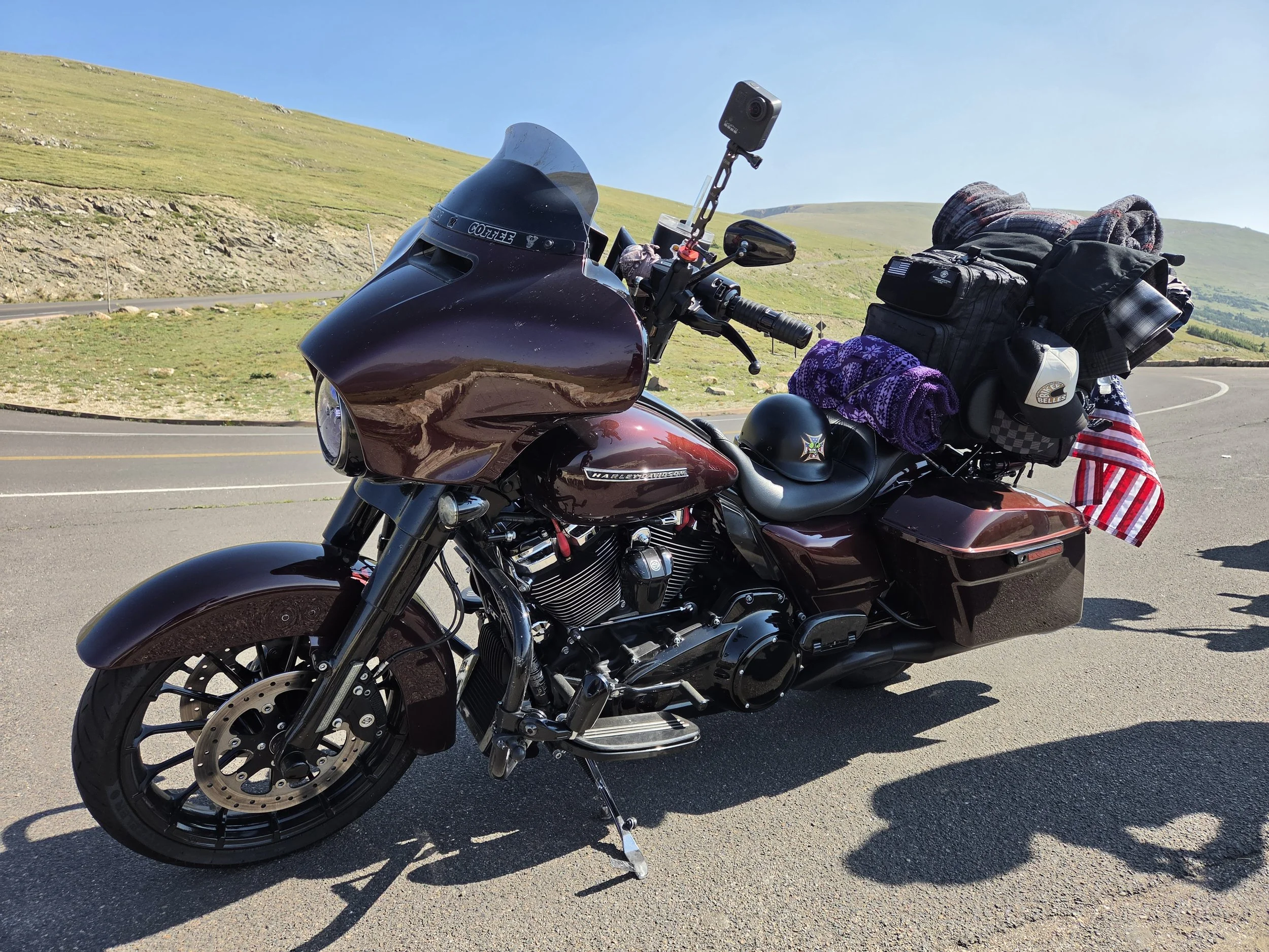 Black Harley-Davidson motorcycle parked on the side of a curving road in a mountainous area with green rolling hills and a clear blue sky in the background. The motorcycle is loaded with bags, rolled-up blankets, and a camera mounted on a hold attach