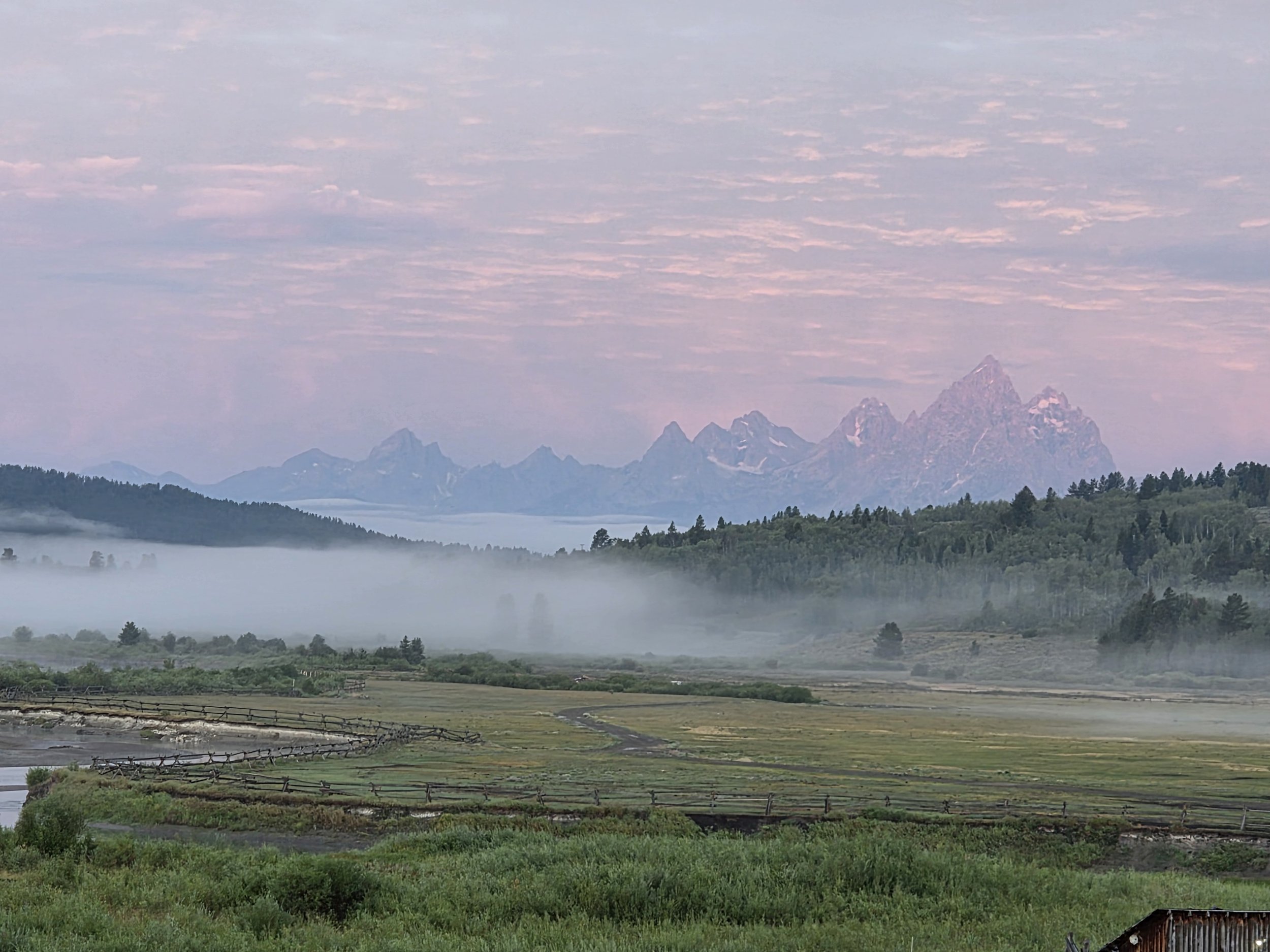 Mountain range with snow-capped peaks in the background, layered with mist and clouds, foreground showing lush green fields and trees, early morning or late evening lighting with soft pinkish hues in the sky.
