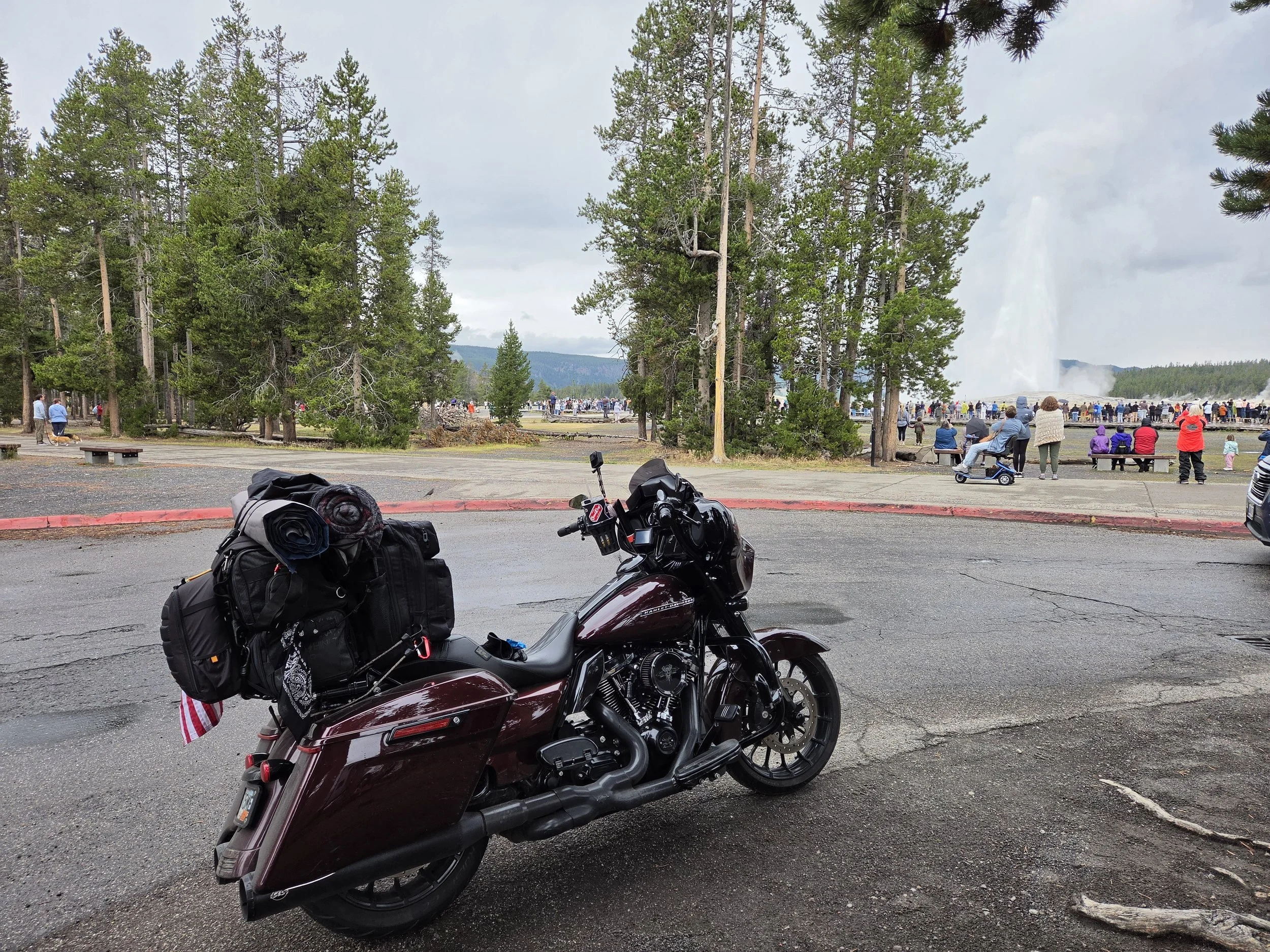 A motorcycle parked in a lot with packed camping gear attached, overlooking a crowd near a geyser erupting in a park surrounded by tall trees and distant mountains.