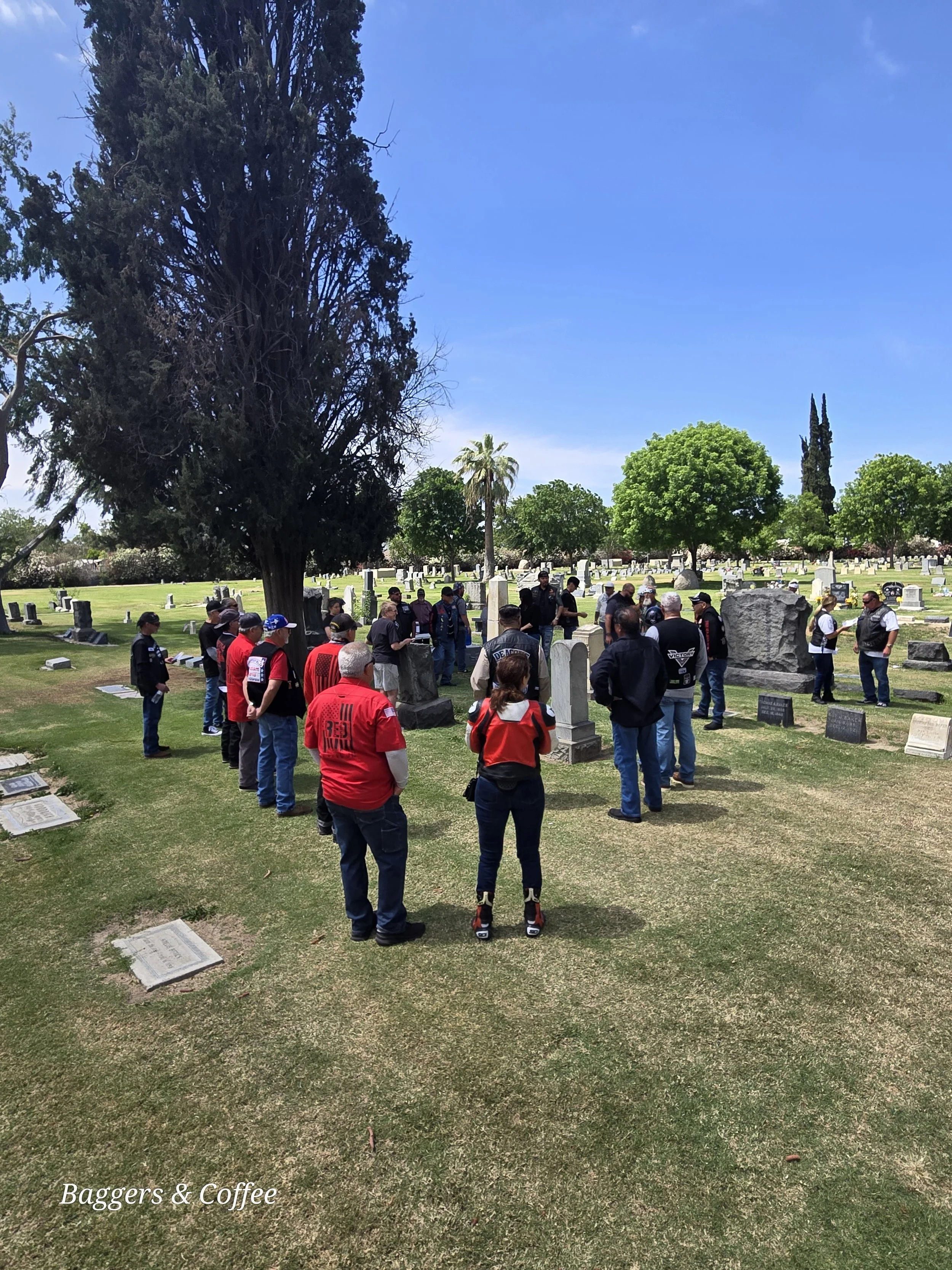 A group of people gathered in a cemetery with gravestones, surrounded by trees and a clear blue sky.