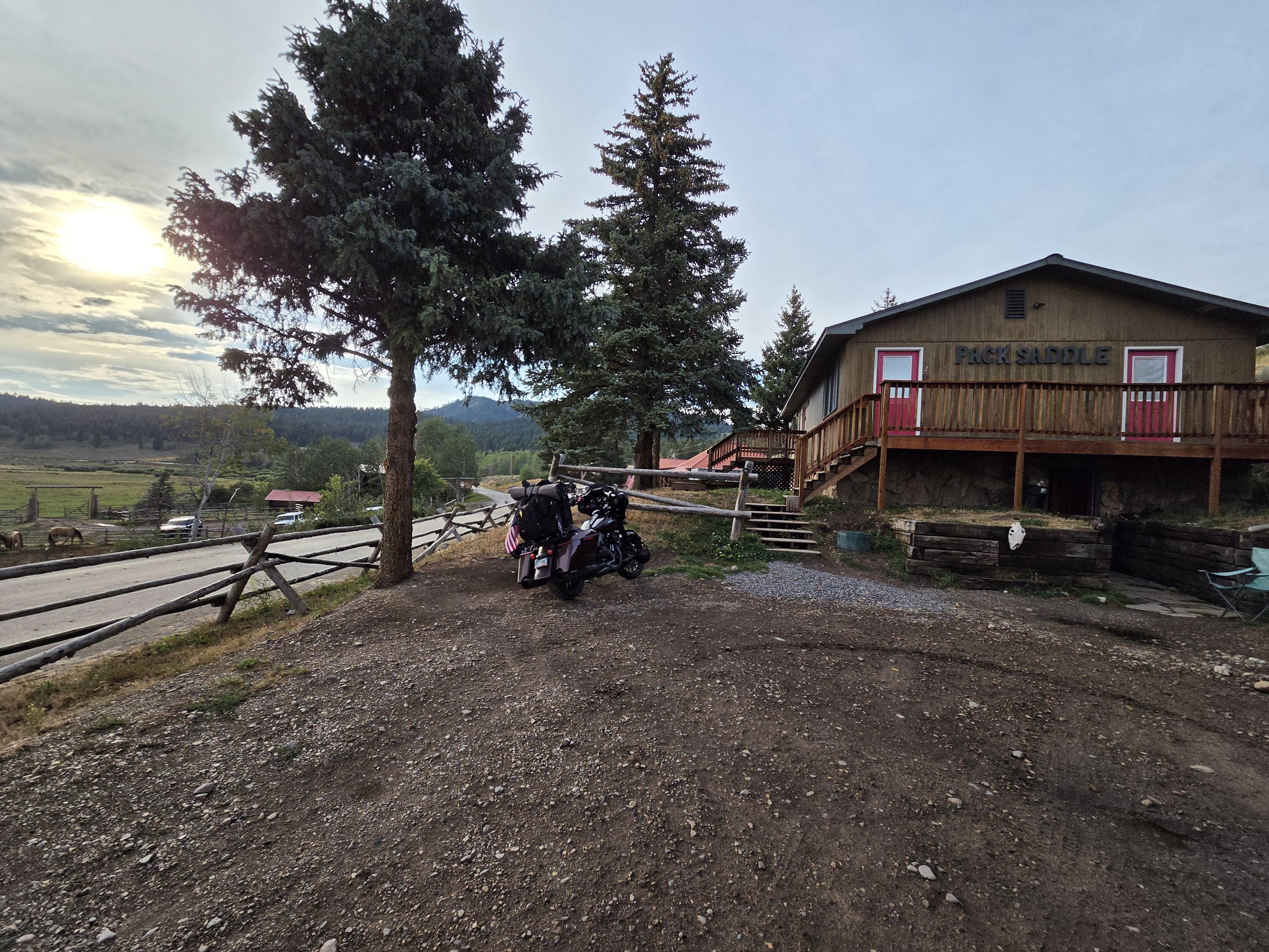 A rustic wooden building labeled 'PICK SADDLE' with a balcony, surrounded by tall pine trees, in a rural landscape during sunset, with a motorcycle and some animals nearby.