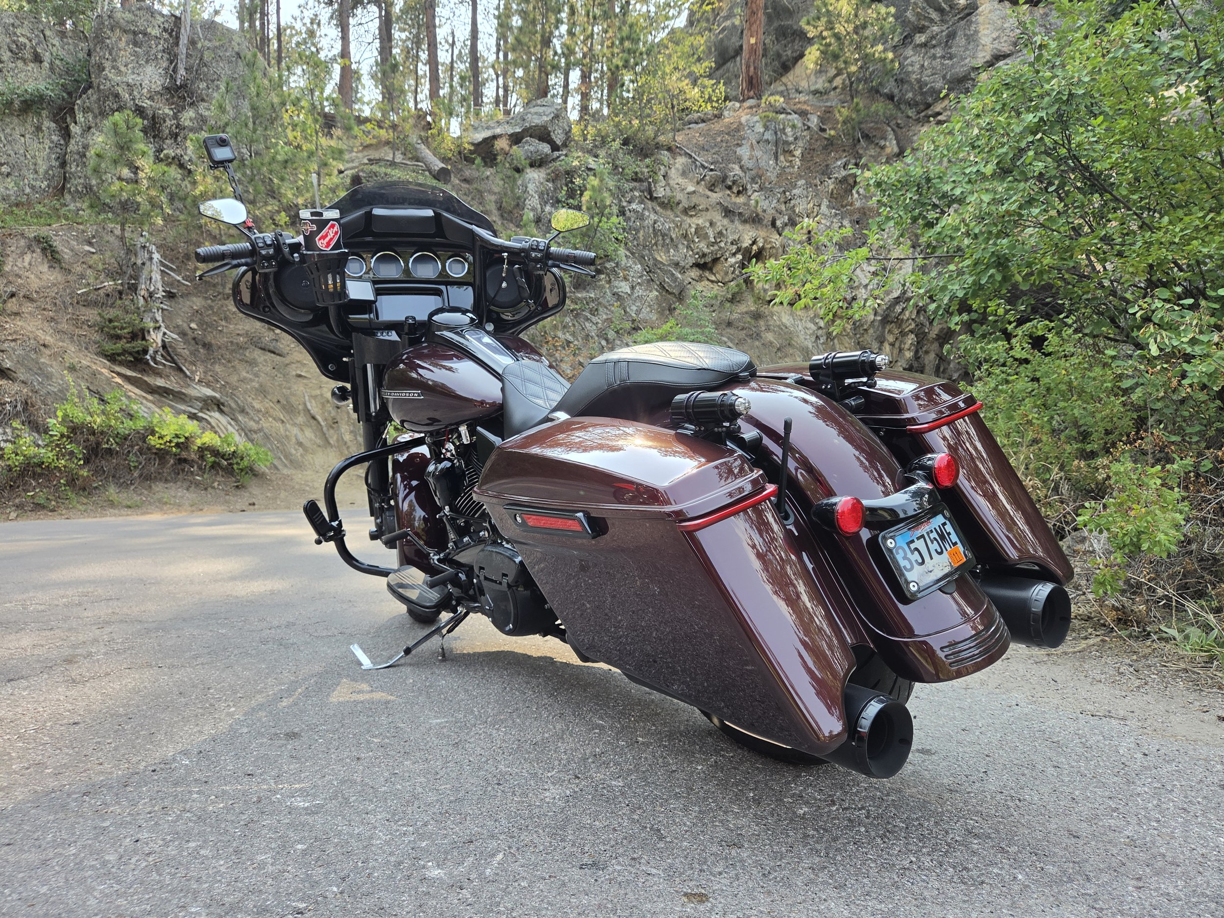 A burgundy Harley-Davidson touring motorcycle parked on the side of a dirt road in a wooded area with pine trees and rocks in the background.