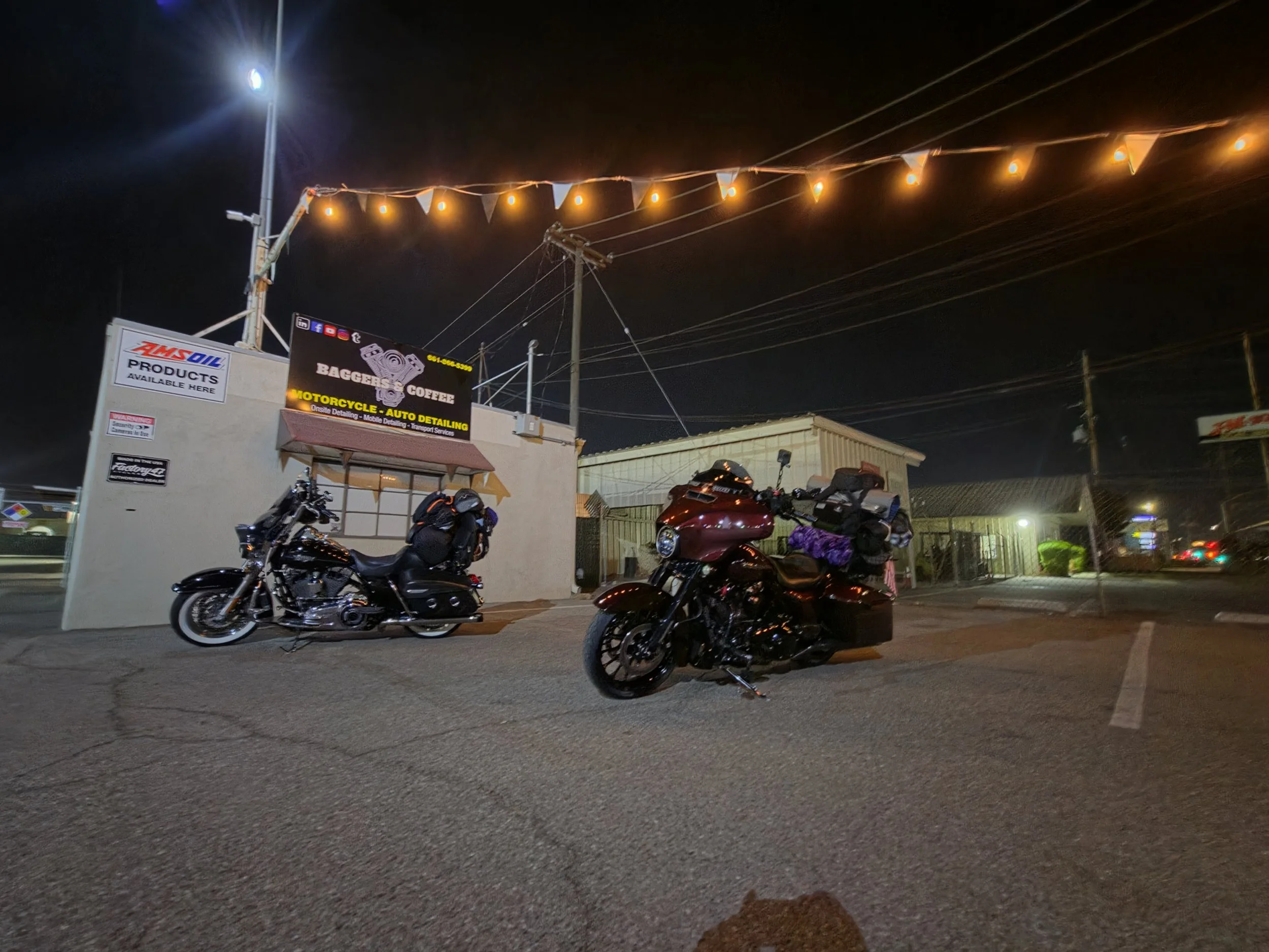 Nighttime scene of a parking lot with motorcycles, a building with signs, and string lights hanging above.