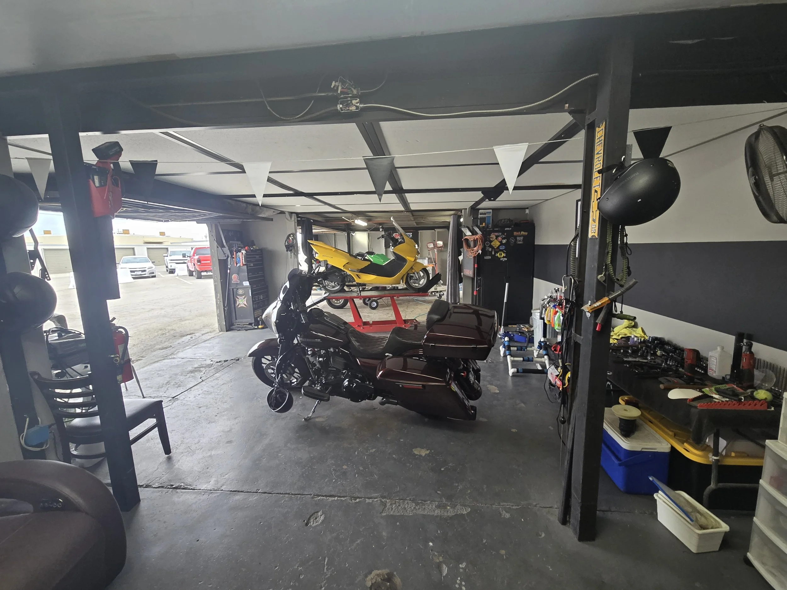 View of a garage containing motorcycles, car parts, and tools, with an open garage door and parked cars outside.
