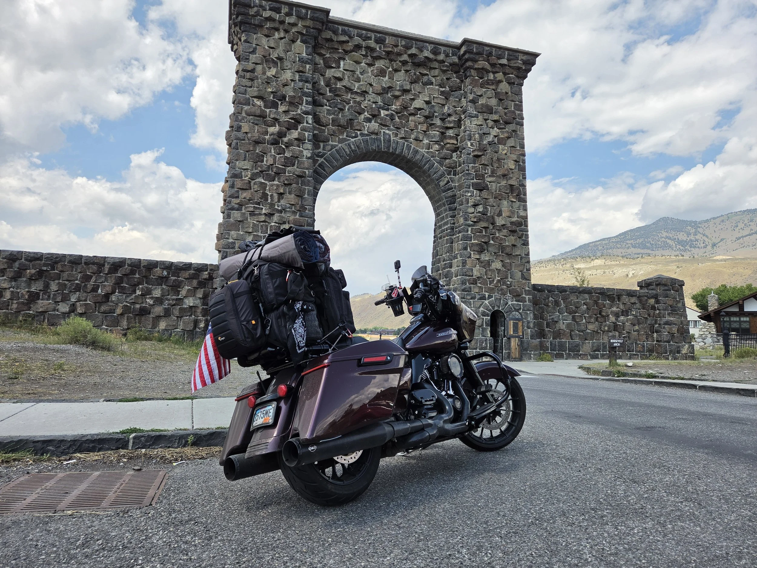 A motorcycle parked on a street with a stone archway structure in the background, with hills and a partly cloudy sky behind it.