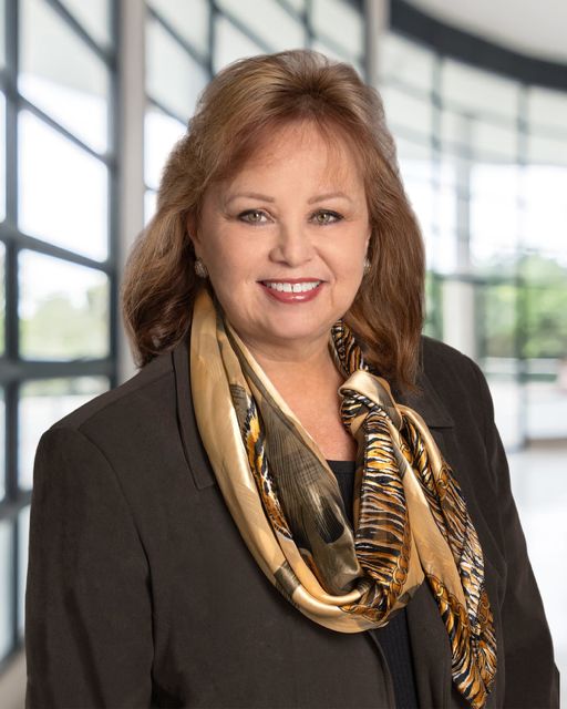 Professional woman with shoulder-length red hair wearing a black blazer and a multicolored silk scarf, inside a modern glass building.