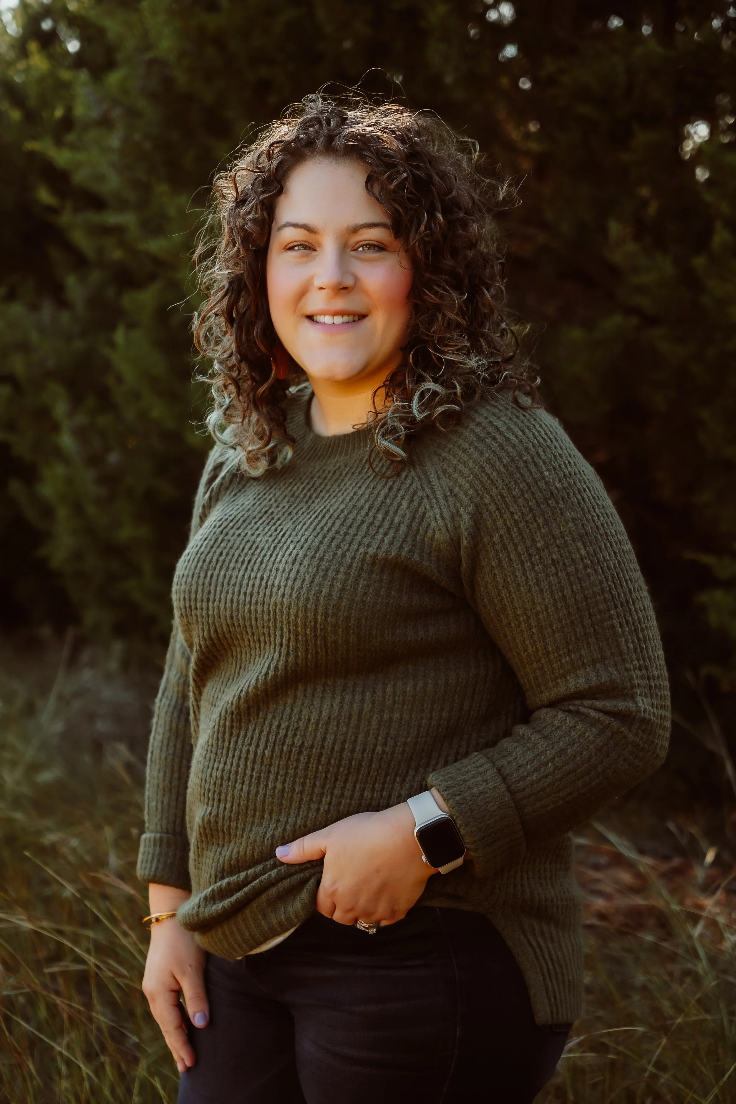 Young woman with curly hair smiling outdoors, wearing a green sweater, black pants, and a smartwatch, standing in front of trees and grass.
