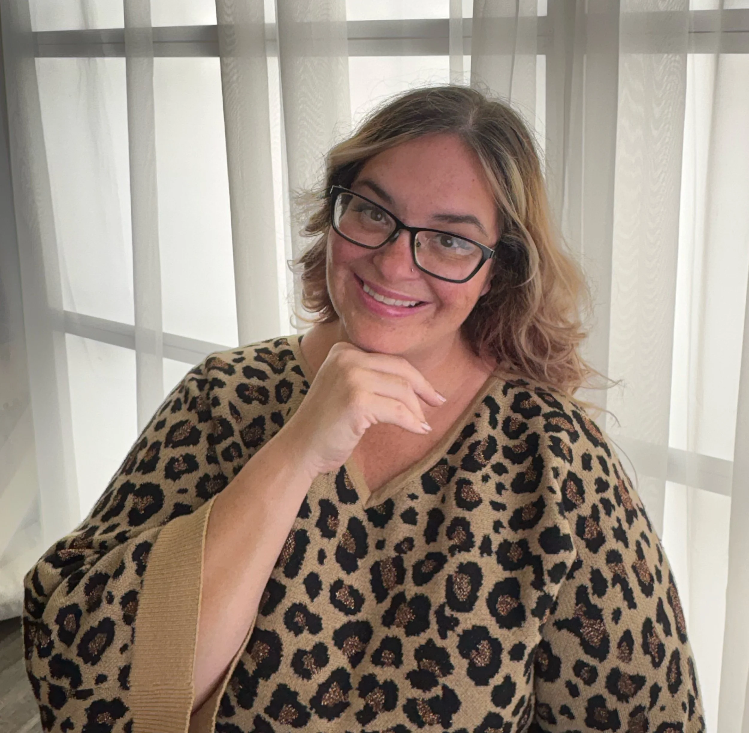 A woman with glasses and wavy blonde hair smiling, wearing a leopard print top, near a window with sheer curtains.