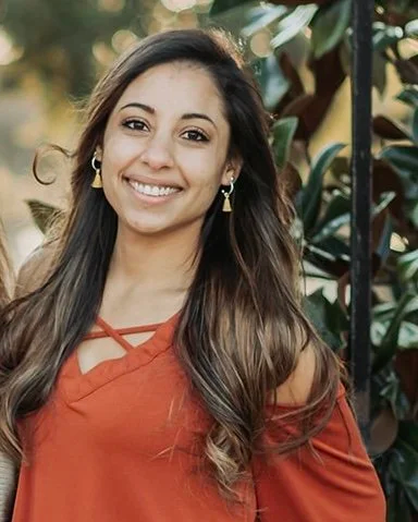 A young woman with long wavy hair smiling outdoors, wearing a rust-colored off-shoulder top and earrings, with greenery in the background.