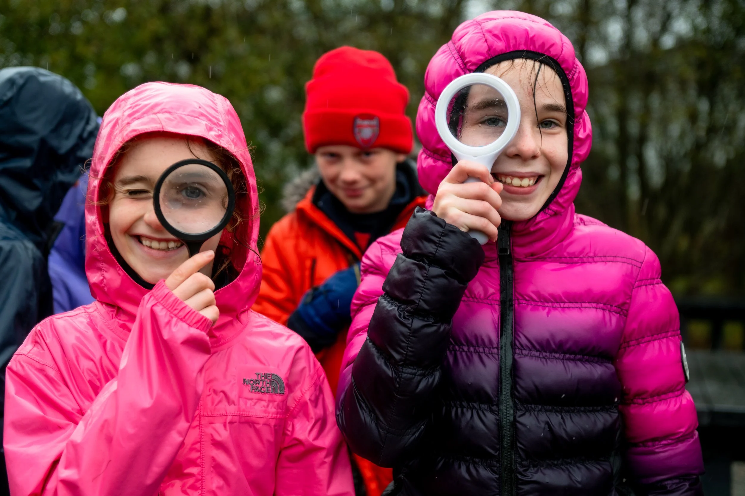 Three children outdoors dressed in colorful rain jackets and hats, with two holding magnifying glasses, exploring or playing in rainy weather.
