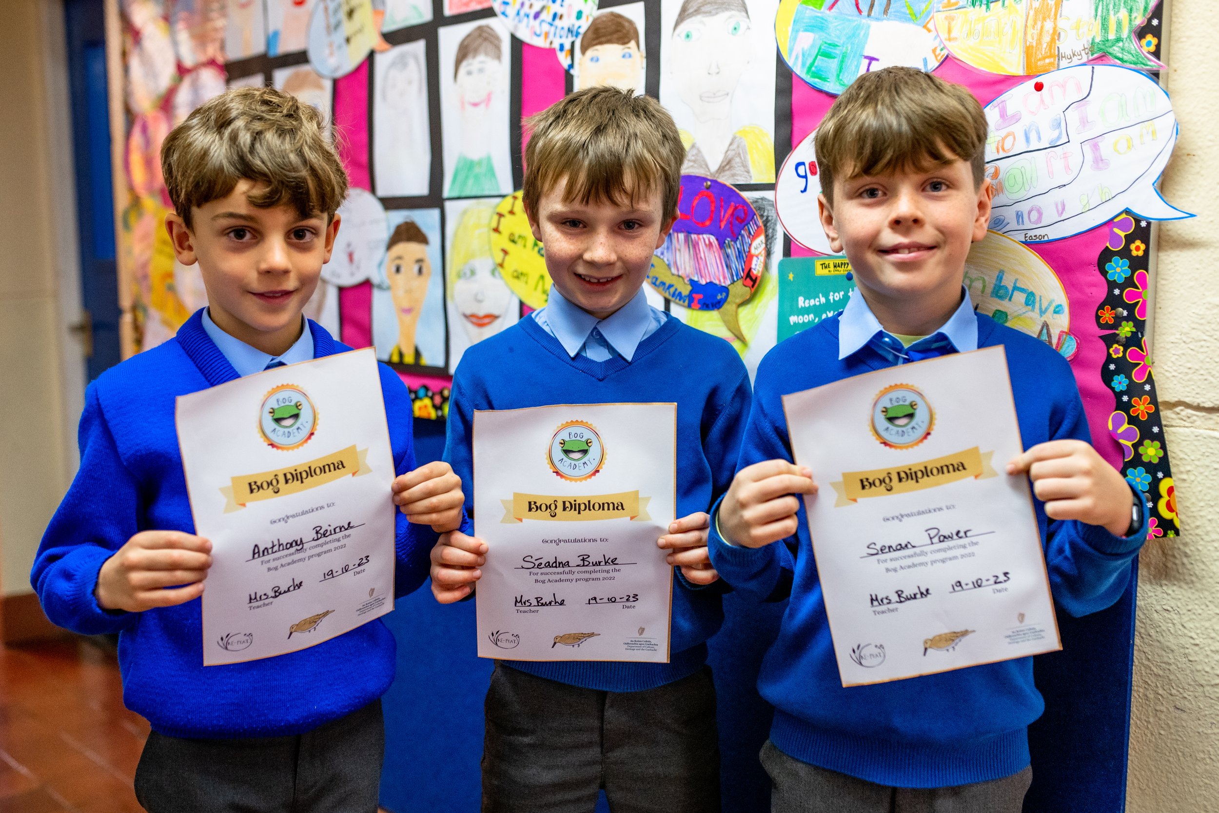 Three young boys in blue school uniforms holding certificates of achievement and smiling at the camera, standing in front of a colorful school bulletin board decorated with student artwork and speech bubbles.