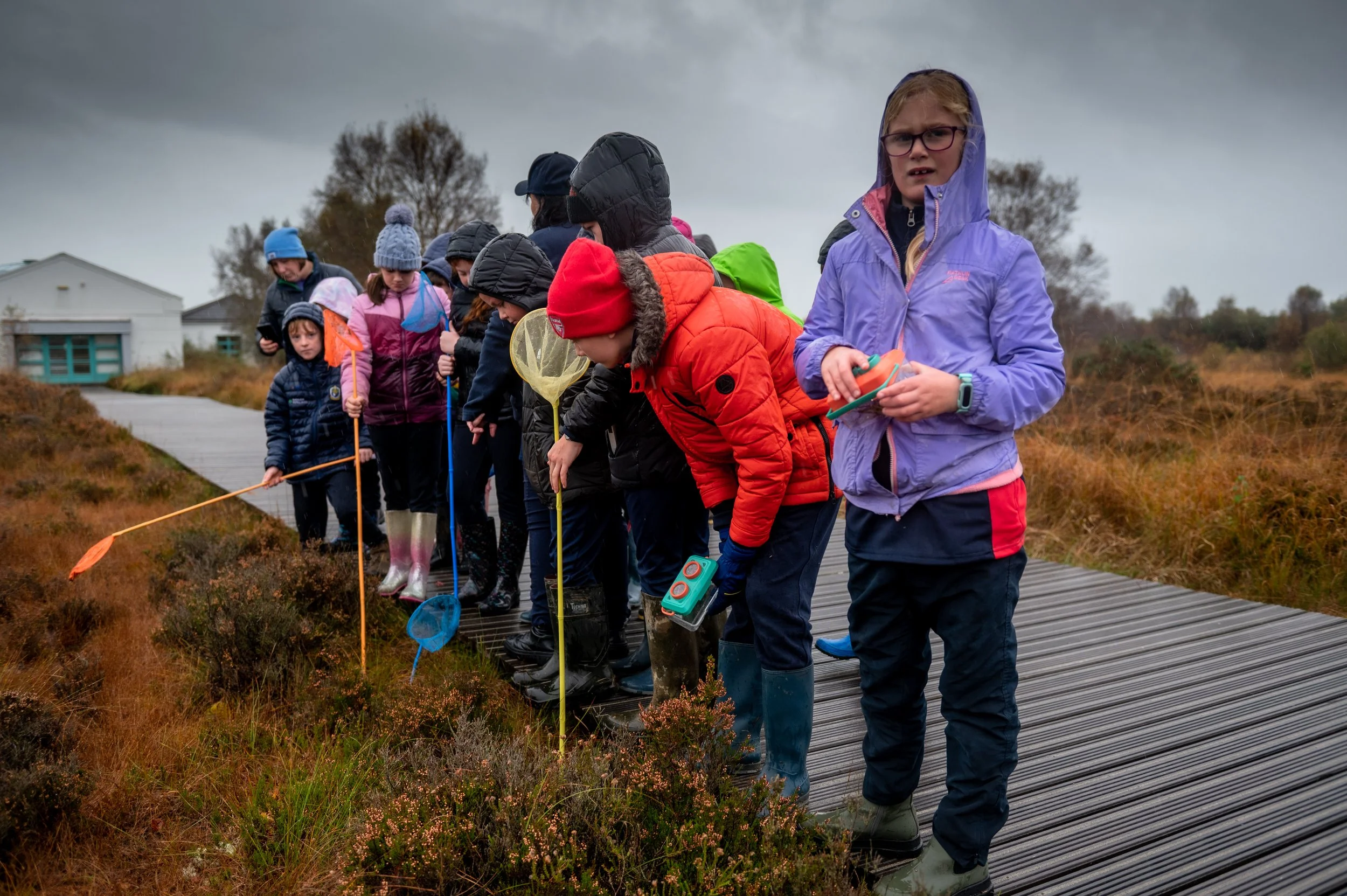 Group of children in waterproof clothing and boots, standing on a boardwalk in a wet, marshy area outdoors on a cloudy day. Some children are holding insect nets, and a girl in a purple jacket and glasses is holding a container.