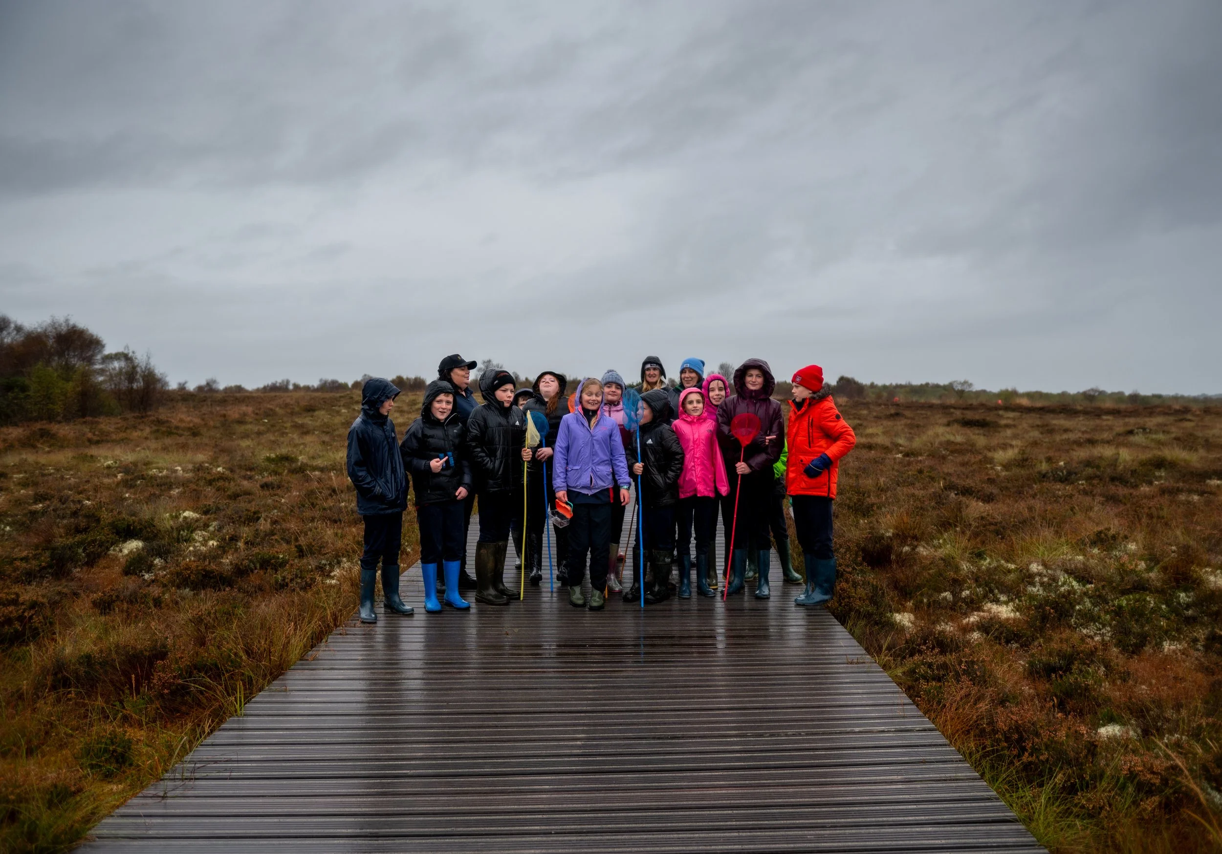 Group of children and adults dressed in rain jackets and boots standing on a wooden boardwalk in an open field on a cloudy day.