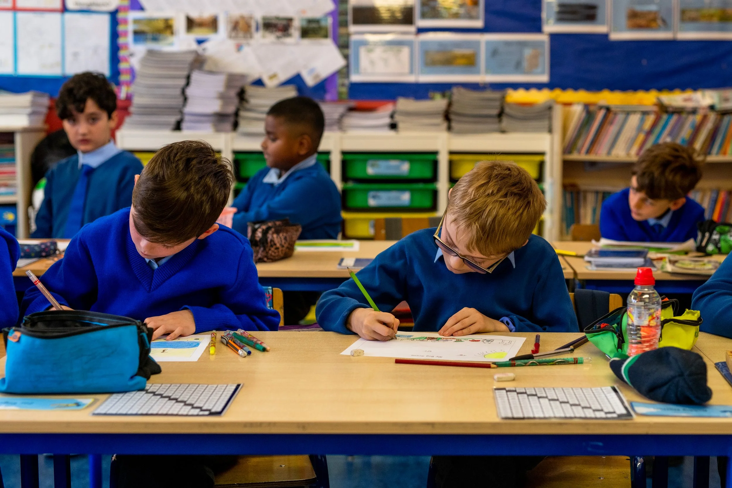 Children sitting at desks in a classroom, focused on drawing or writing, with books and supplies on the desks and shelves in the background.